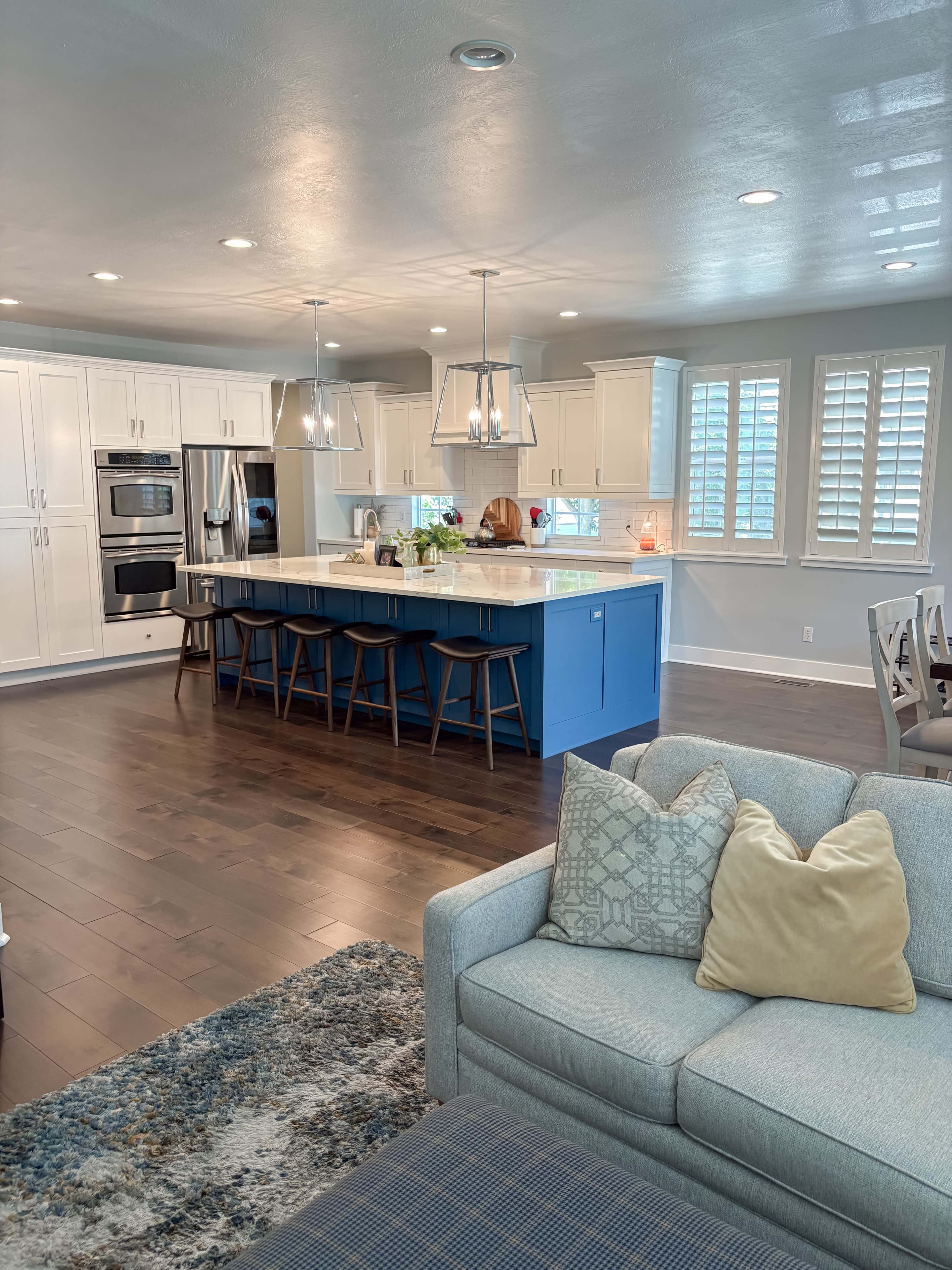 A modern kitchen features white cabinets, a blue island with seating, and hardwood floors, while a comfortable living area with a gray sofa and decorative pillows is visible nearby.