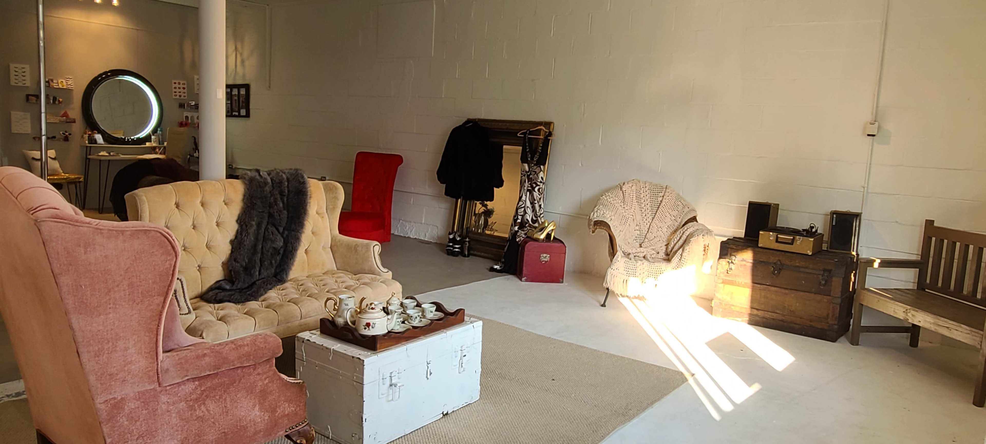 The image shows a brightly lit, minimalist room featuring assorted seating, including a pink armchair, a red chair, and a wooden bench, along with a coffee table and a vintage trunk.