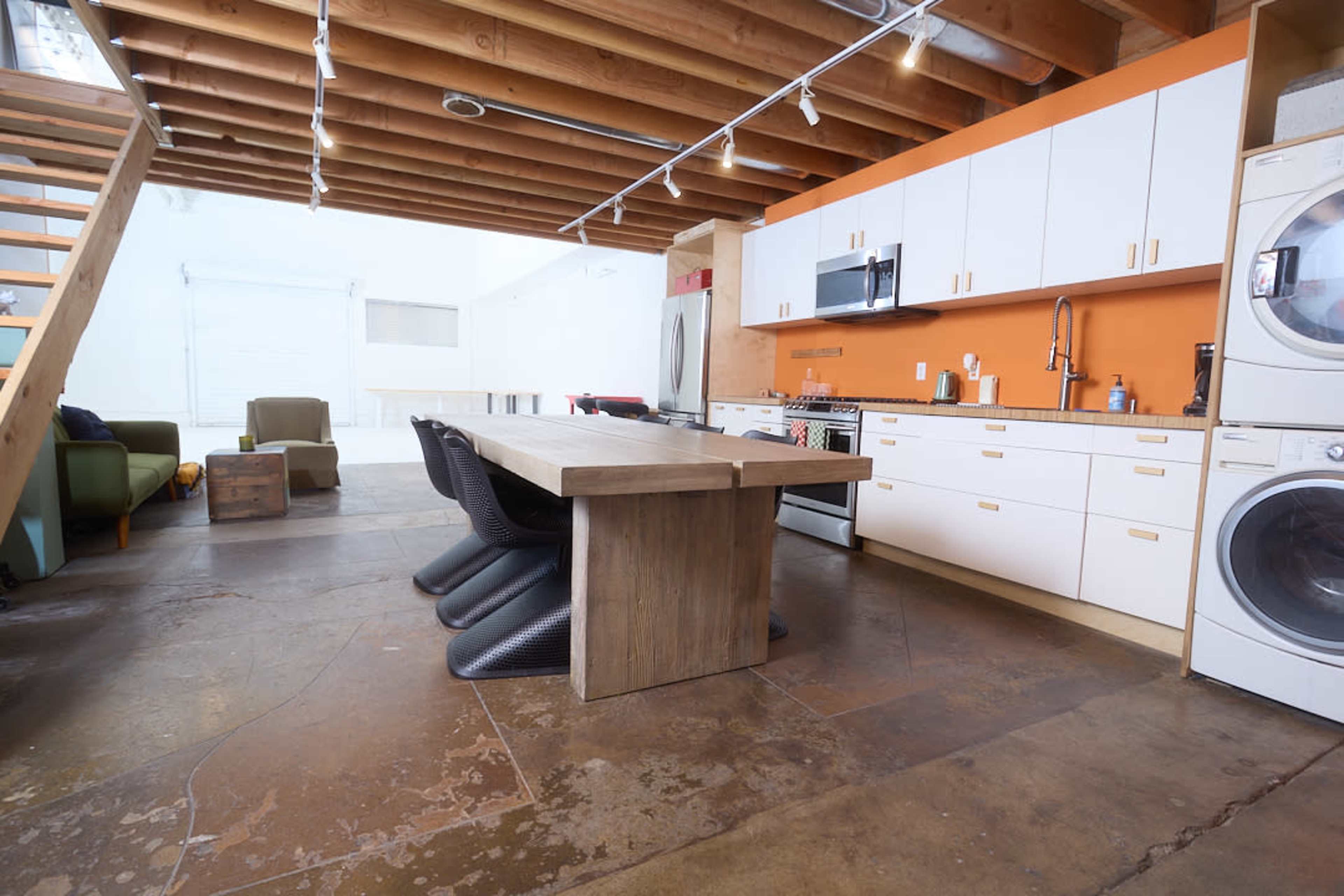 The image shows an open kitchen and dining area with industrial elements, featuring a large wooden table, modern appliances, and a staircase against a wall with exposed beams.