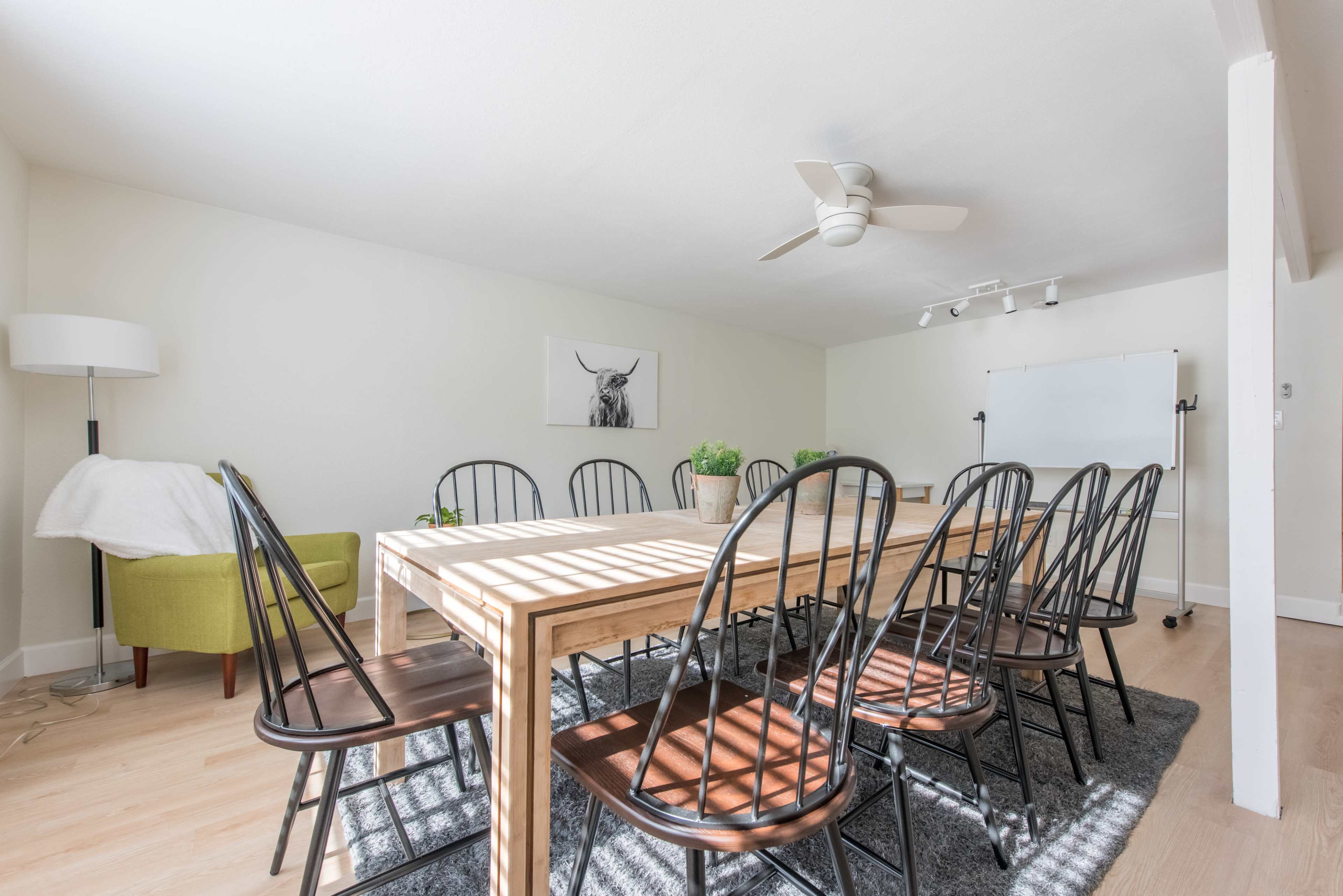 A bright meeting room features a long wooden table surrounded by black chairs, with a green armchair in one corner and a whiteboard nearby.