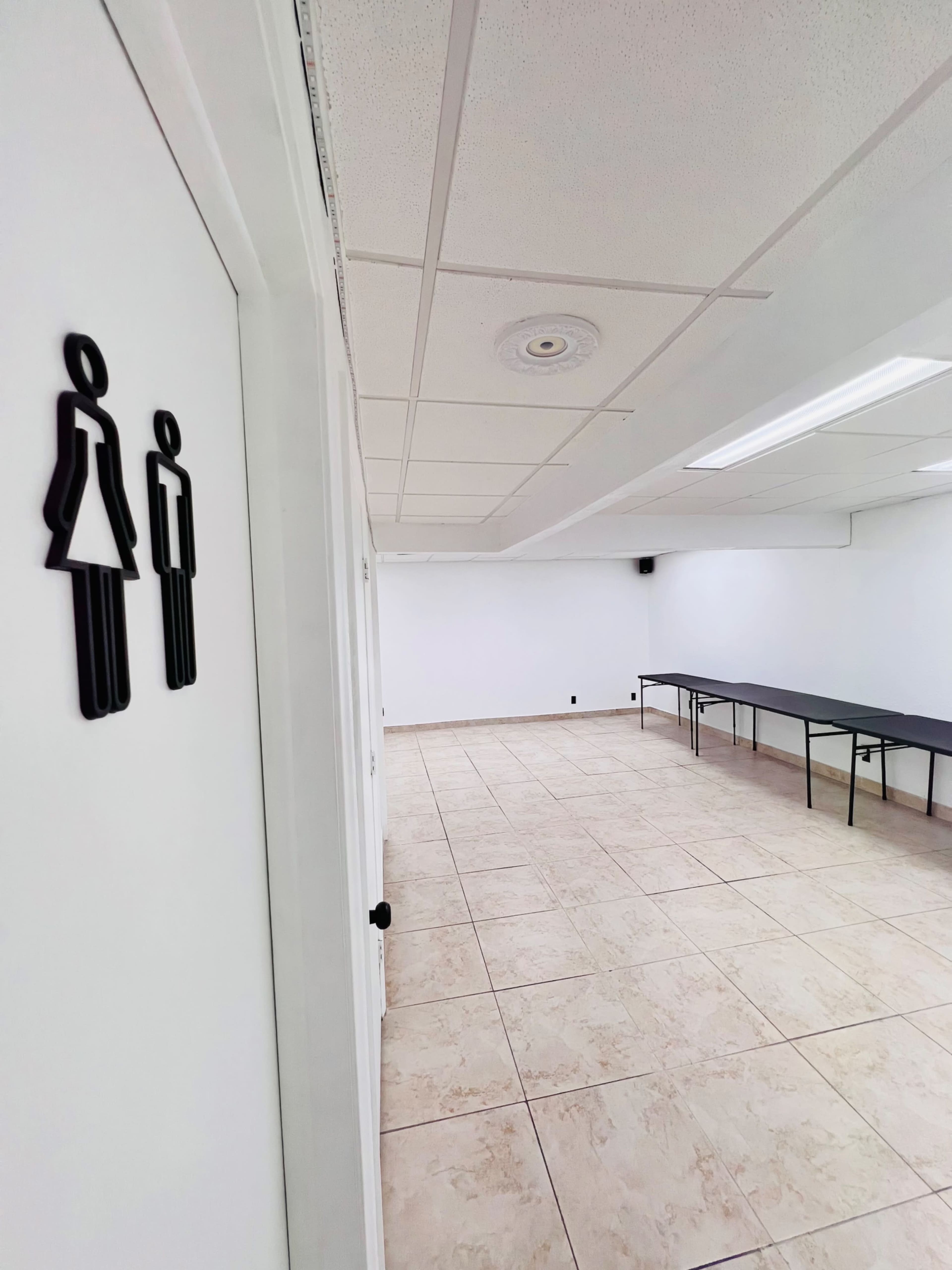 The image shows a hallway leading to restrooms with signs for male and female, and a long table set up in an empty, well-lit room.