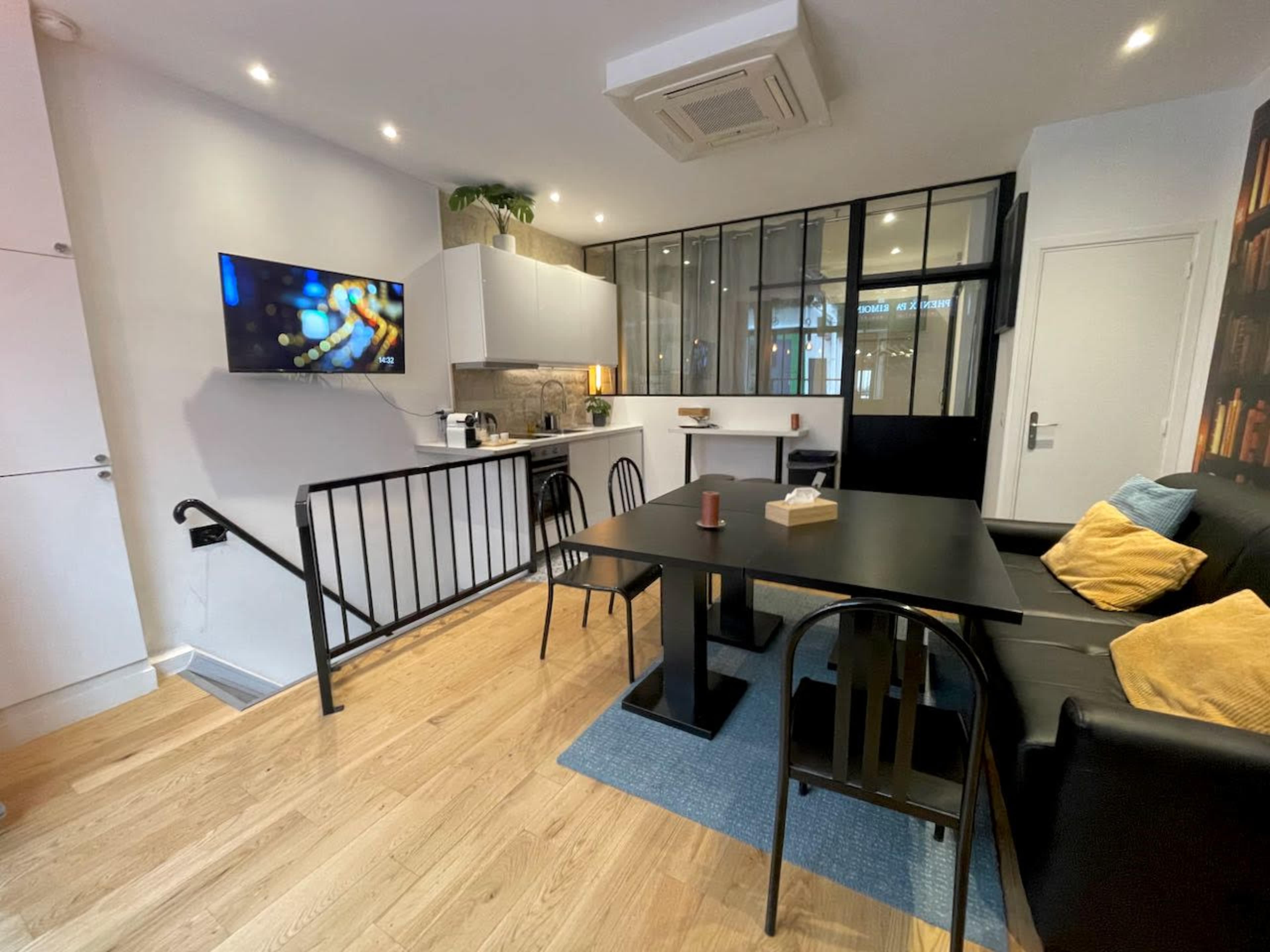 A modern living space featuring a black sofa, a dining table, a kitchenette with a TV mounted on the wall, and a glass partition separating different areas.