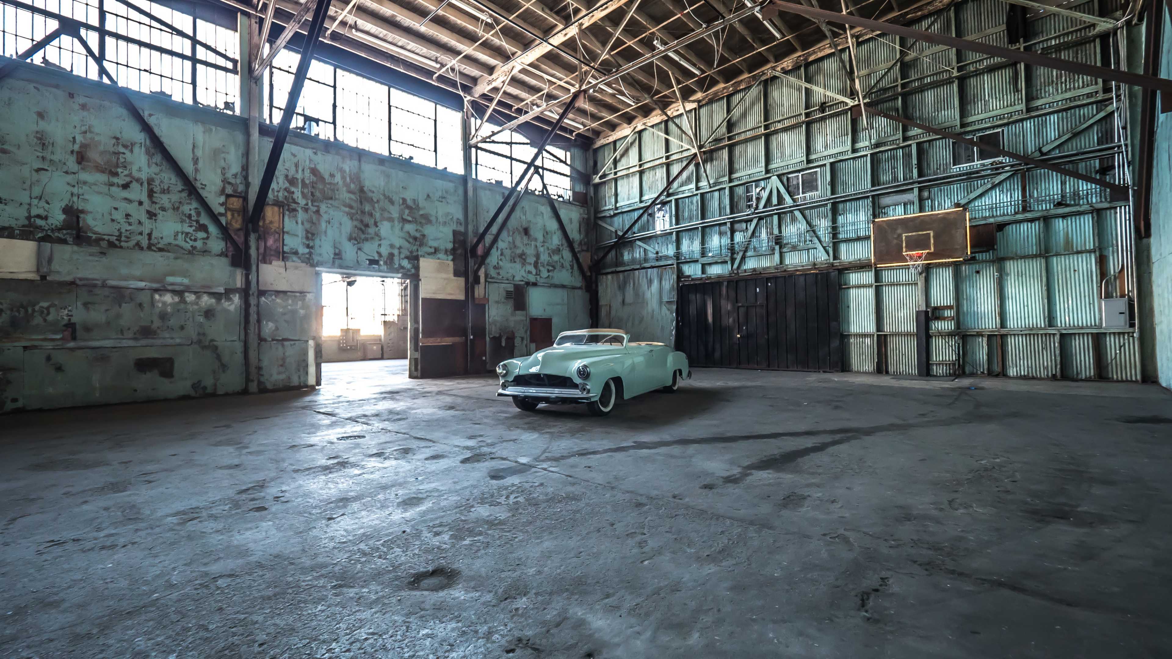 An old turquoise convertible car is parked in the center of a large, empty industrial warehouse with peeling walls and tall windows.