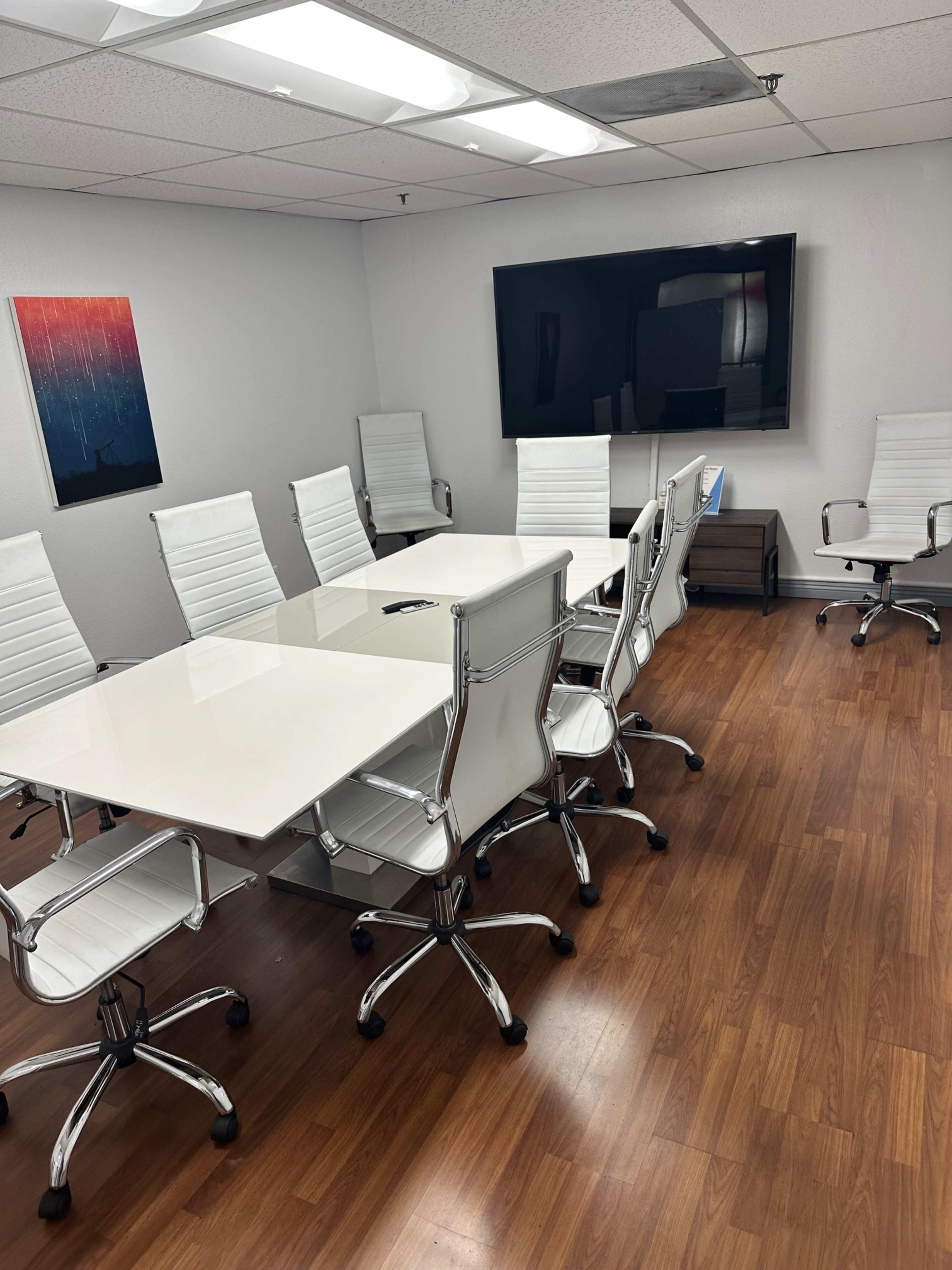 The image shows a modern conference room with a large white table, several white chairs, and a wall-mounted television.