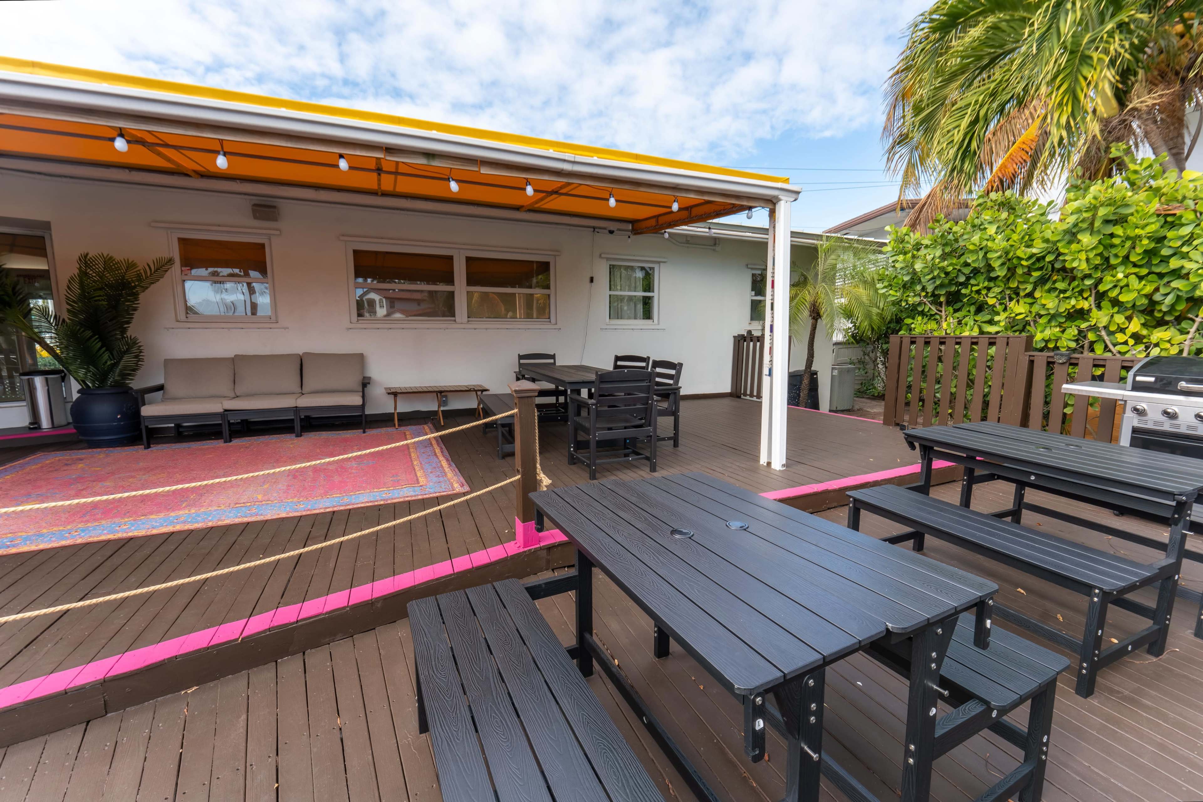 The image shows a patio area with black outdoor furniture, a large colorful rug, and greenery in the background.