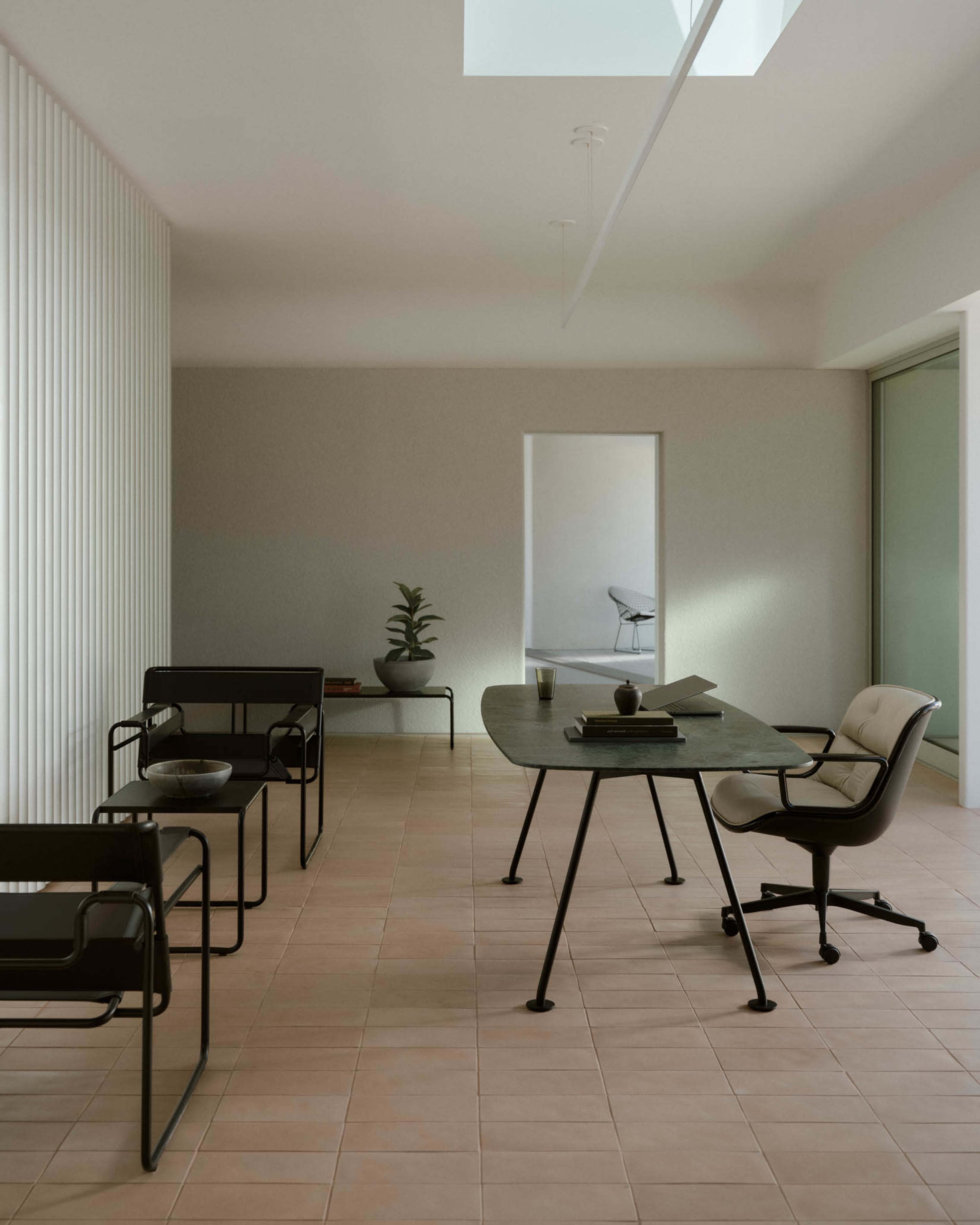 The image depicts a minimalist office space featuring a large table, a rolling chair, two modern black chairs, and a potted plant, with natural light streaming through a skylight.