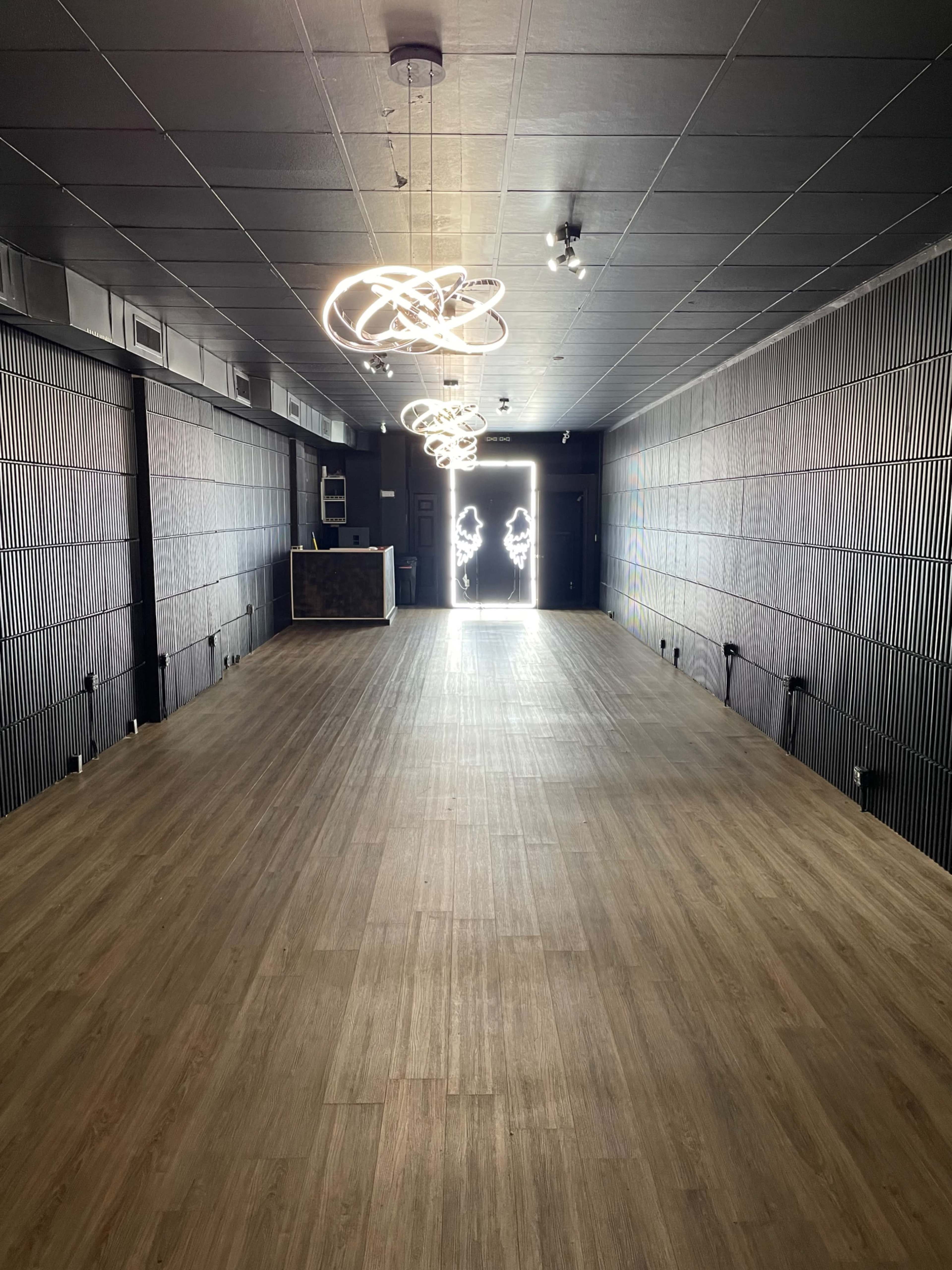 The image shows an empty interior space with wooden flooring, illuminated by modern light fixtures, leading to a bright exit at the far end.