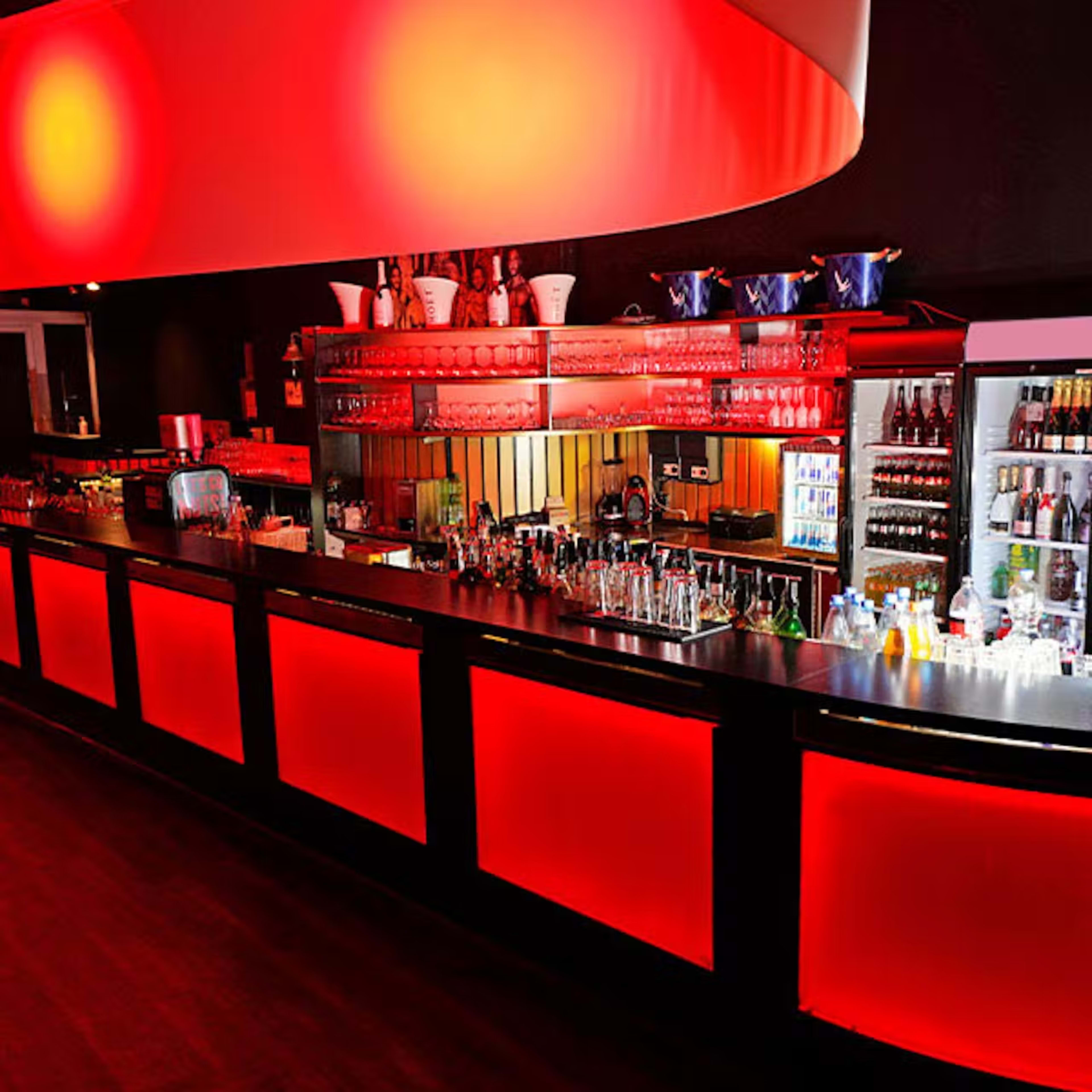 The image shows a sleek bar illuminated by red lighting, showcasing various bottles and glassware on the shelves.
