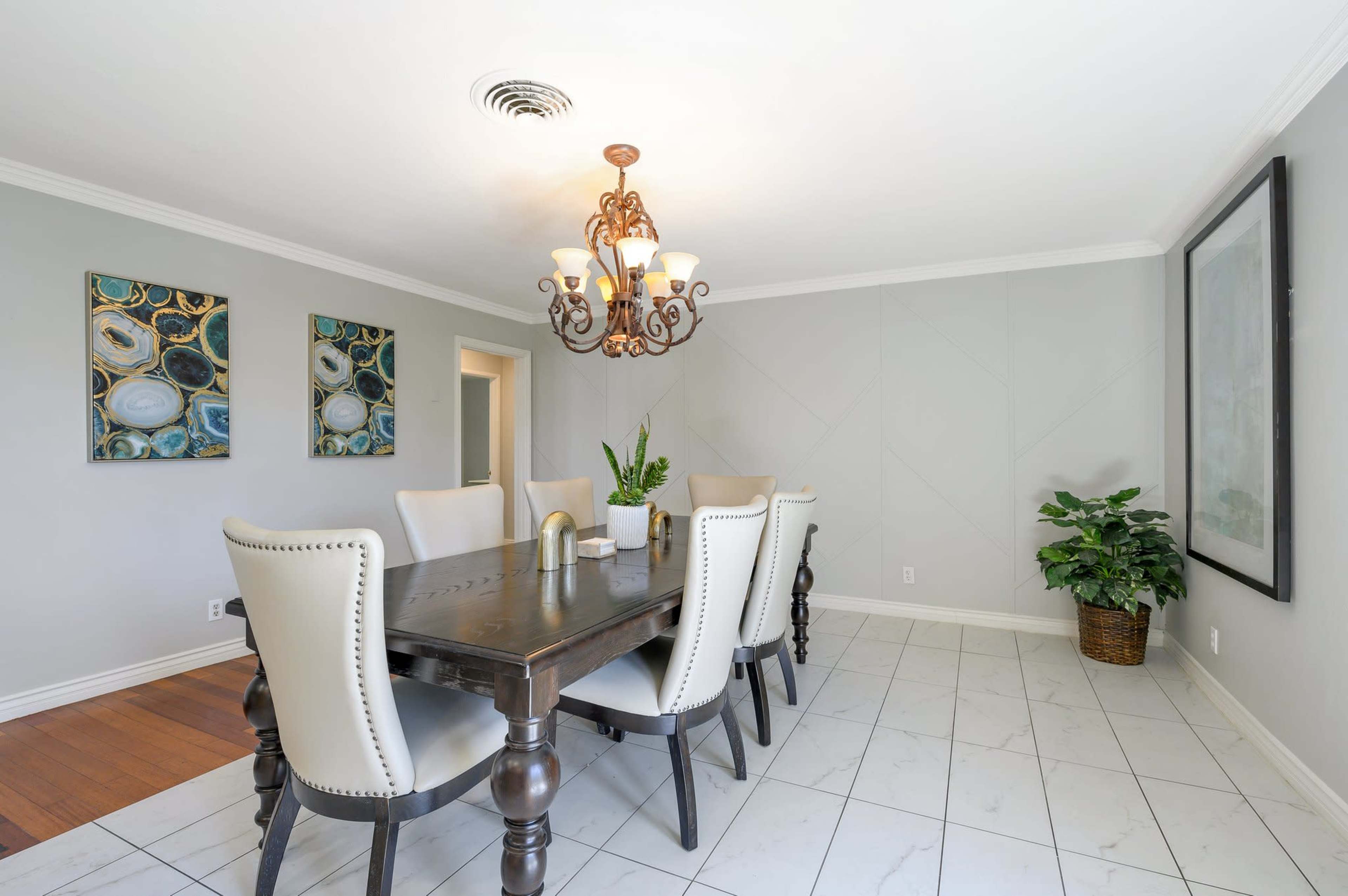 A dining room features a large wooden table surrounded by upholstered chairs, illuminated by a chandelier, with two decorative paintings on the walls and a potted plant in the corner.
