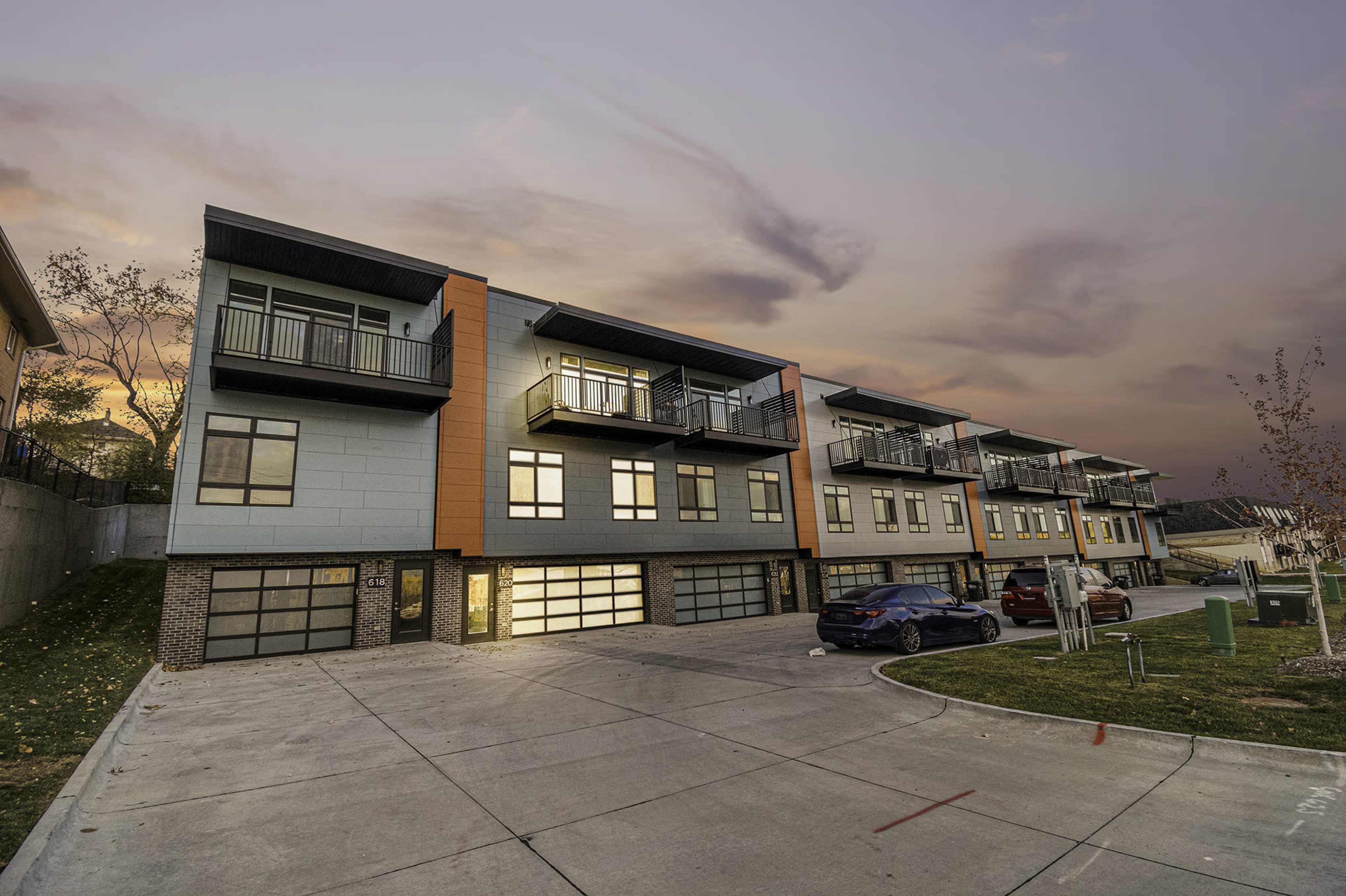 A row of modern townhouses with multiple balconies is situated on a sloped driveway at dusk.