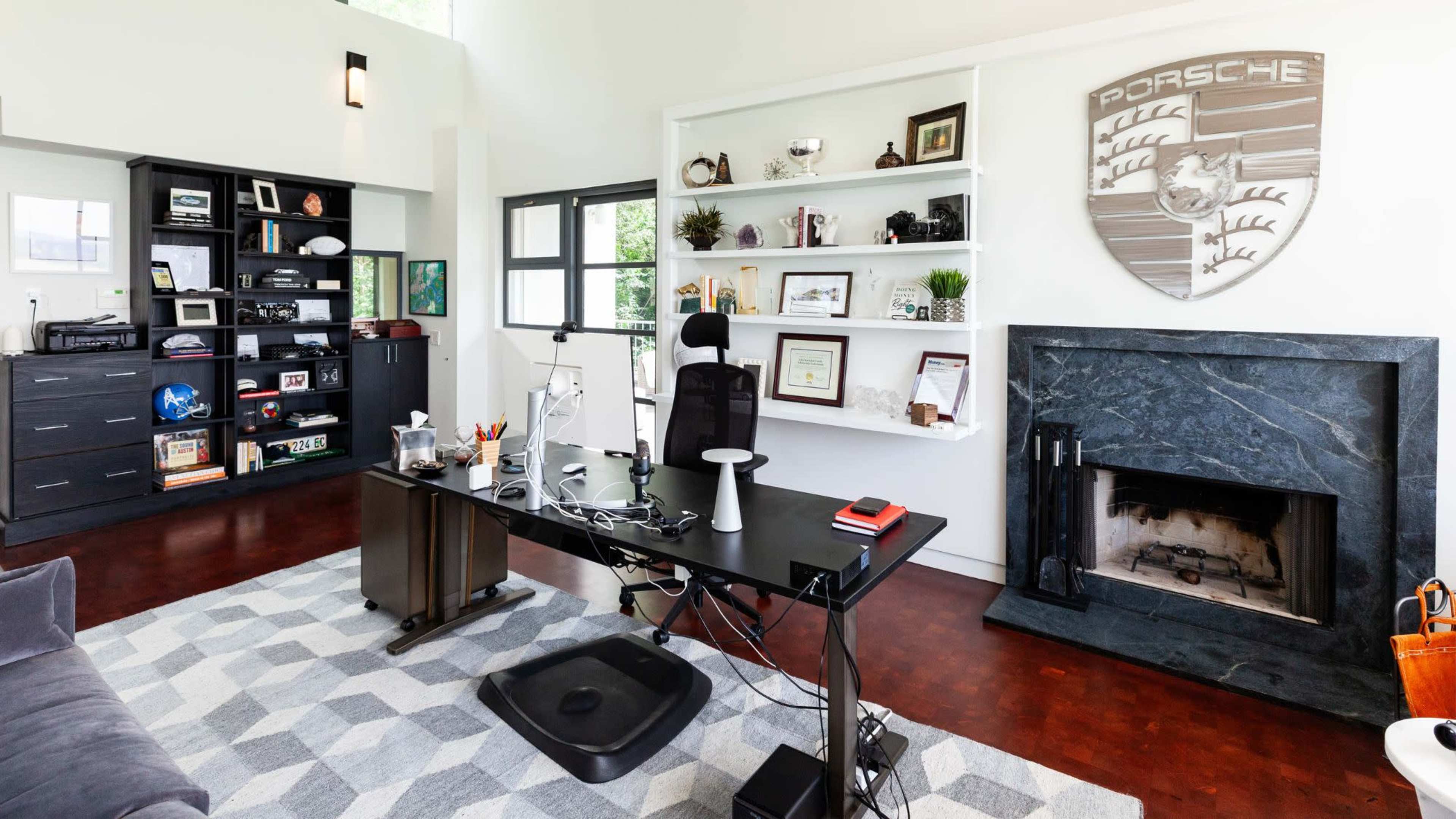 The image shows a modern office with a dark desk, ergonomic chair, and a stone fireplace, featuring a large Porsche crest on the wall and shelves filled with various items.