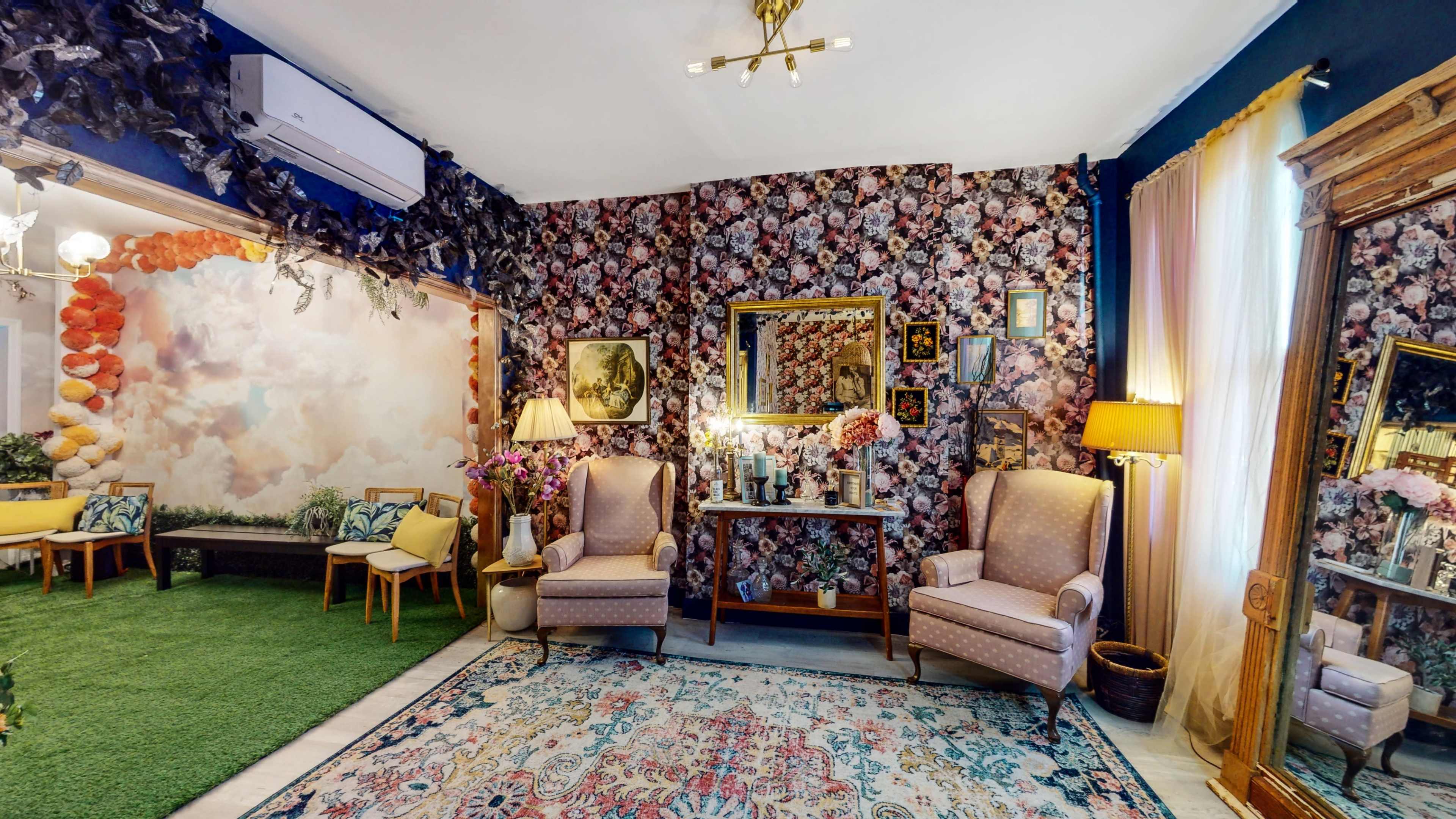 The image depicts a stylish living room featuring floral wallpaper, two upholstered chairs, a decorative mirror, and a patterned area rug.
