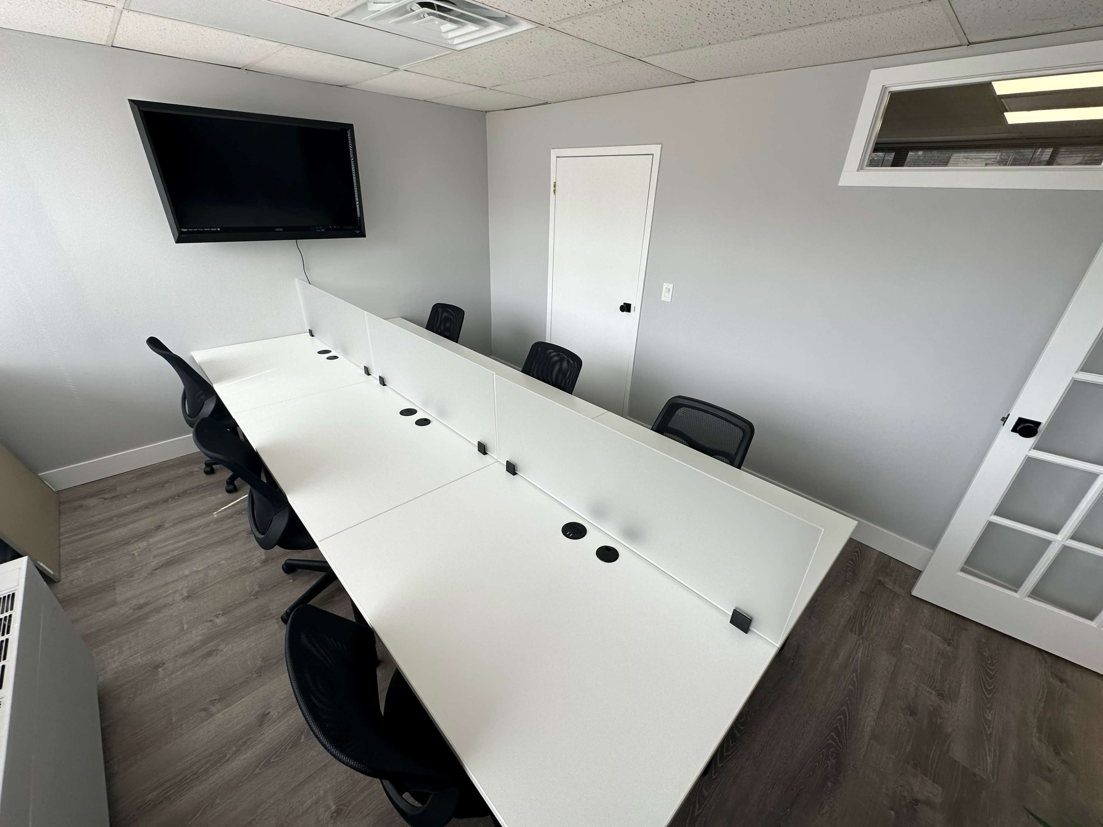 A modern conference room with a long white table, several black chairs, a wall-mounted television, and a door leading to another space.