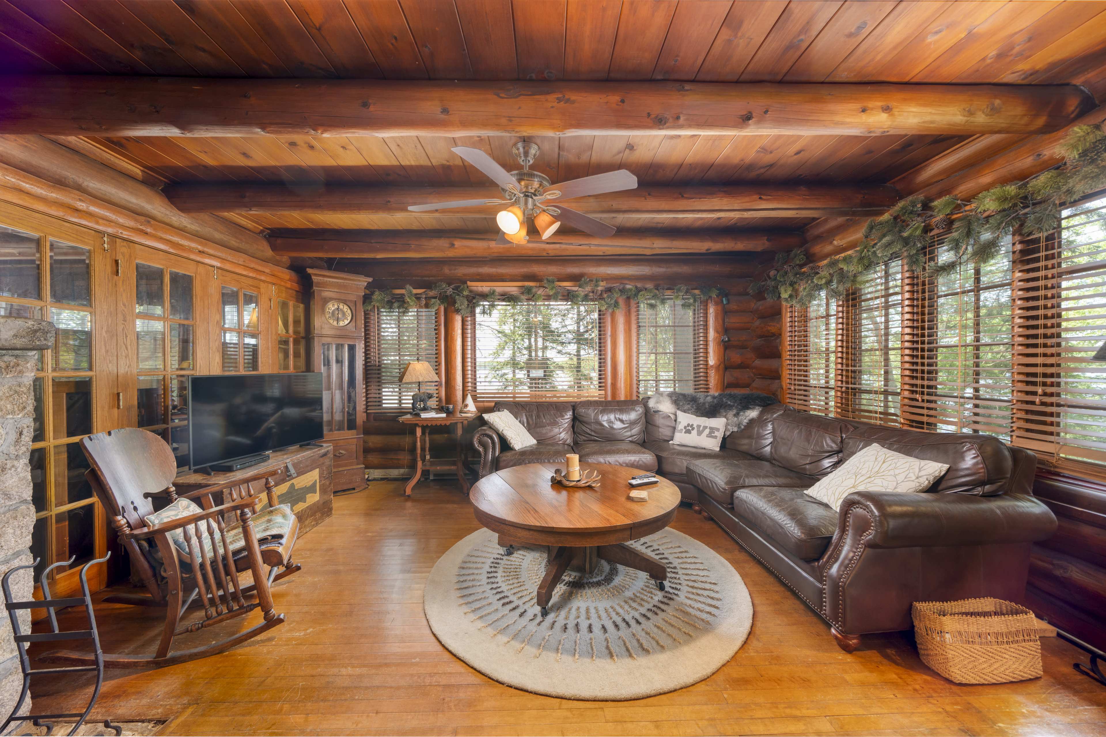 A cozy log cabin living room features a leather sectional sofa, a wooden coffee table, and large windows with blinds.