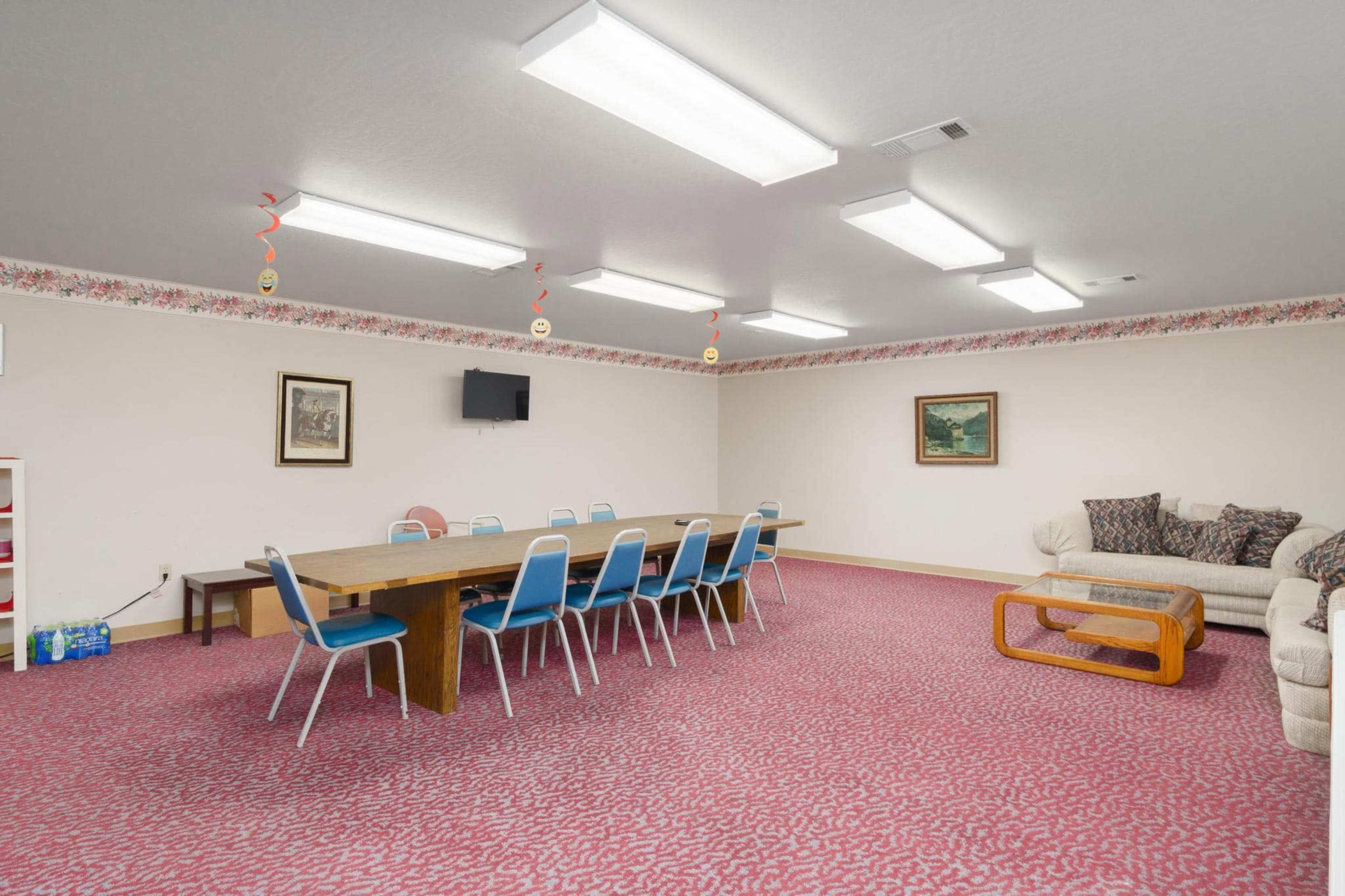 The image shows a well-lit meeting room with a long wooden table surrounded by blue chairs, decorated with floral wallpaper and a couch in one corner.