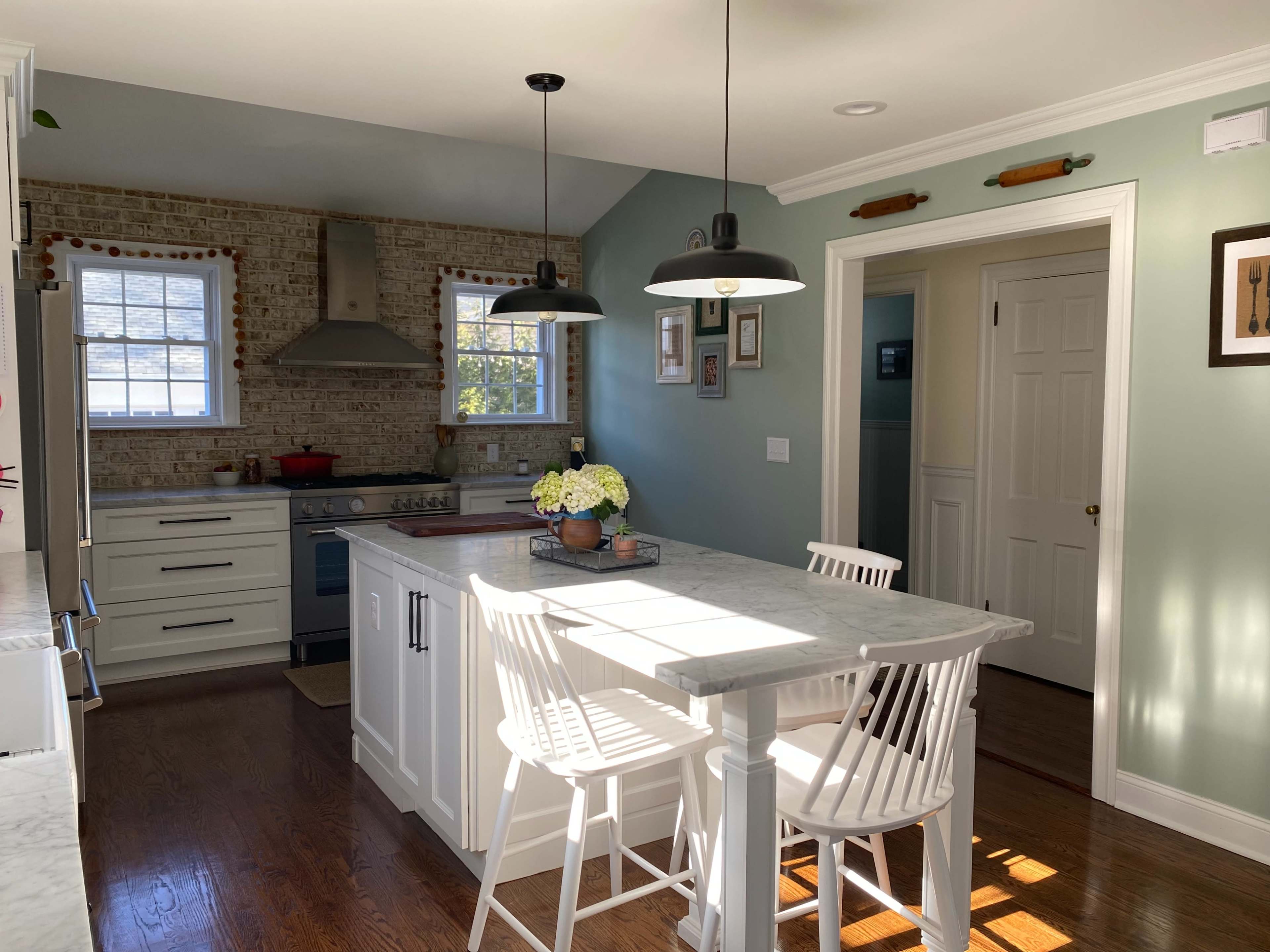 A bright kitchen features a marble island with three white stools, stainless steel appliances, and exposed brick walls.