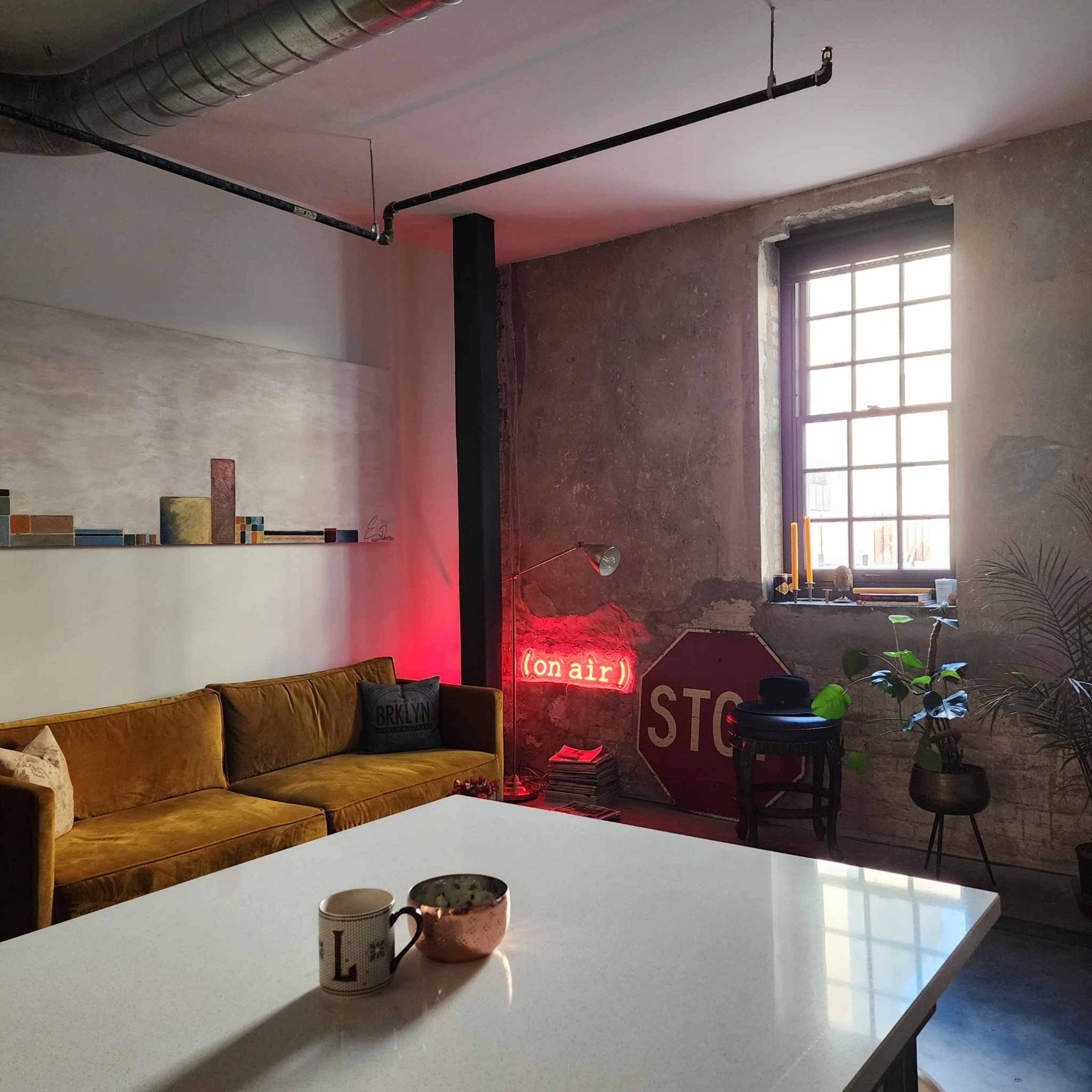 A modern living space features a brown sofa, a large window, a neon "(on air)" sign, and a vintage stop sign against a textured wall.