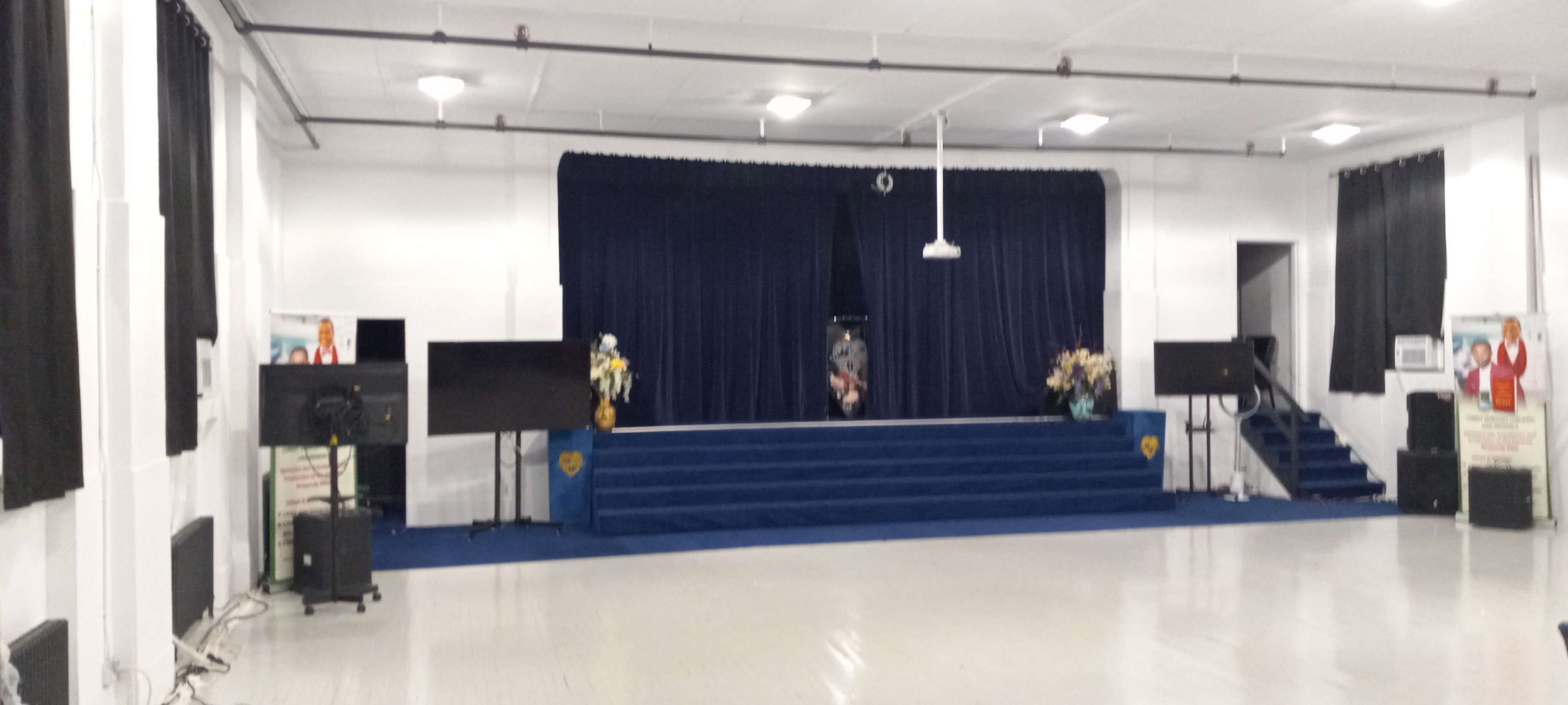 A stage with a blue curtain and steps, flanked by two large screens, in a well-lit hall with white walls and dark curtains.