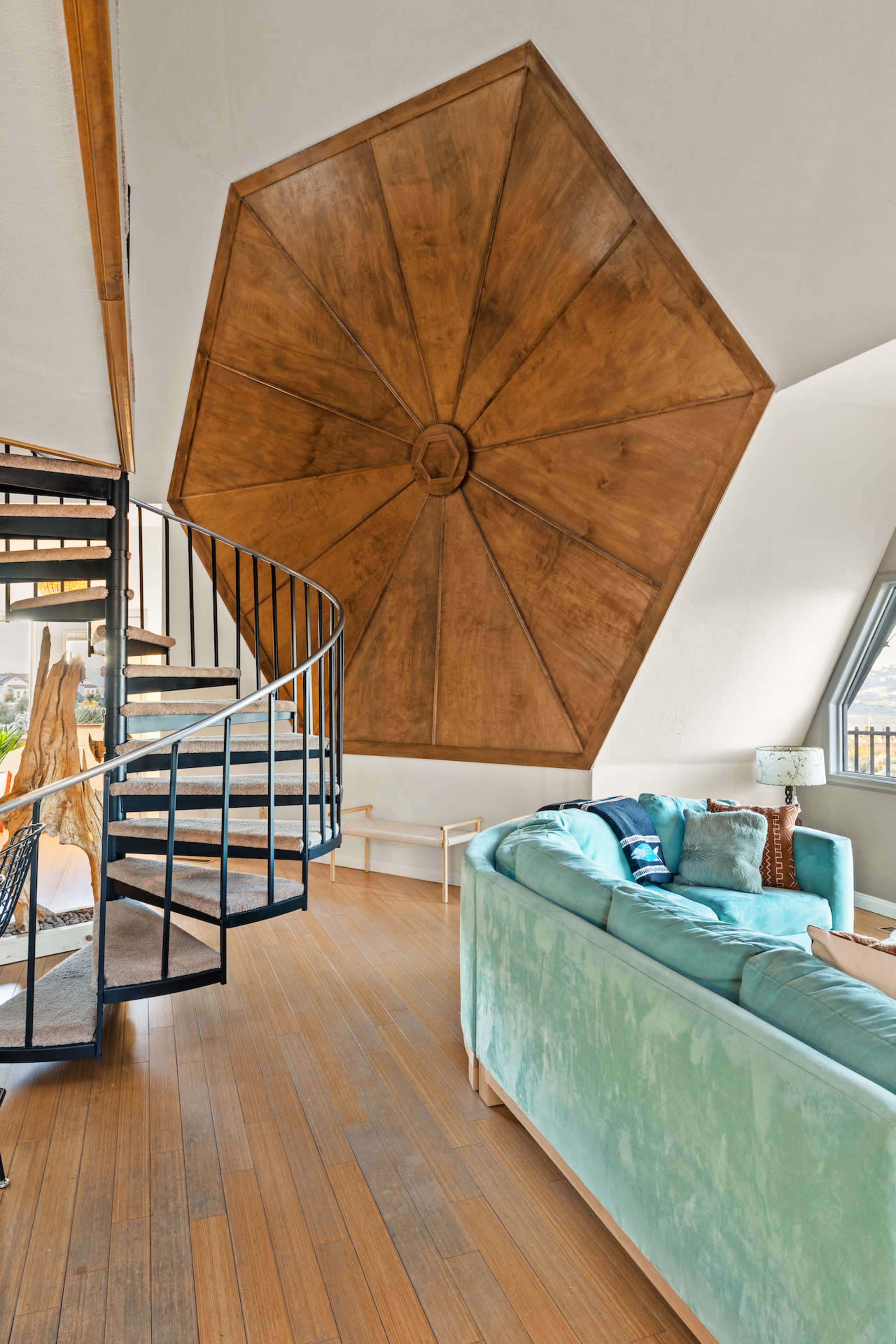 The image shows a modern interior featuring a spiral staircase leading to an upper level, with a large octagonal wooden ceiling above a light-colored sofa.