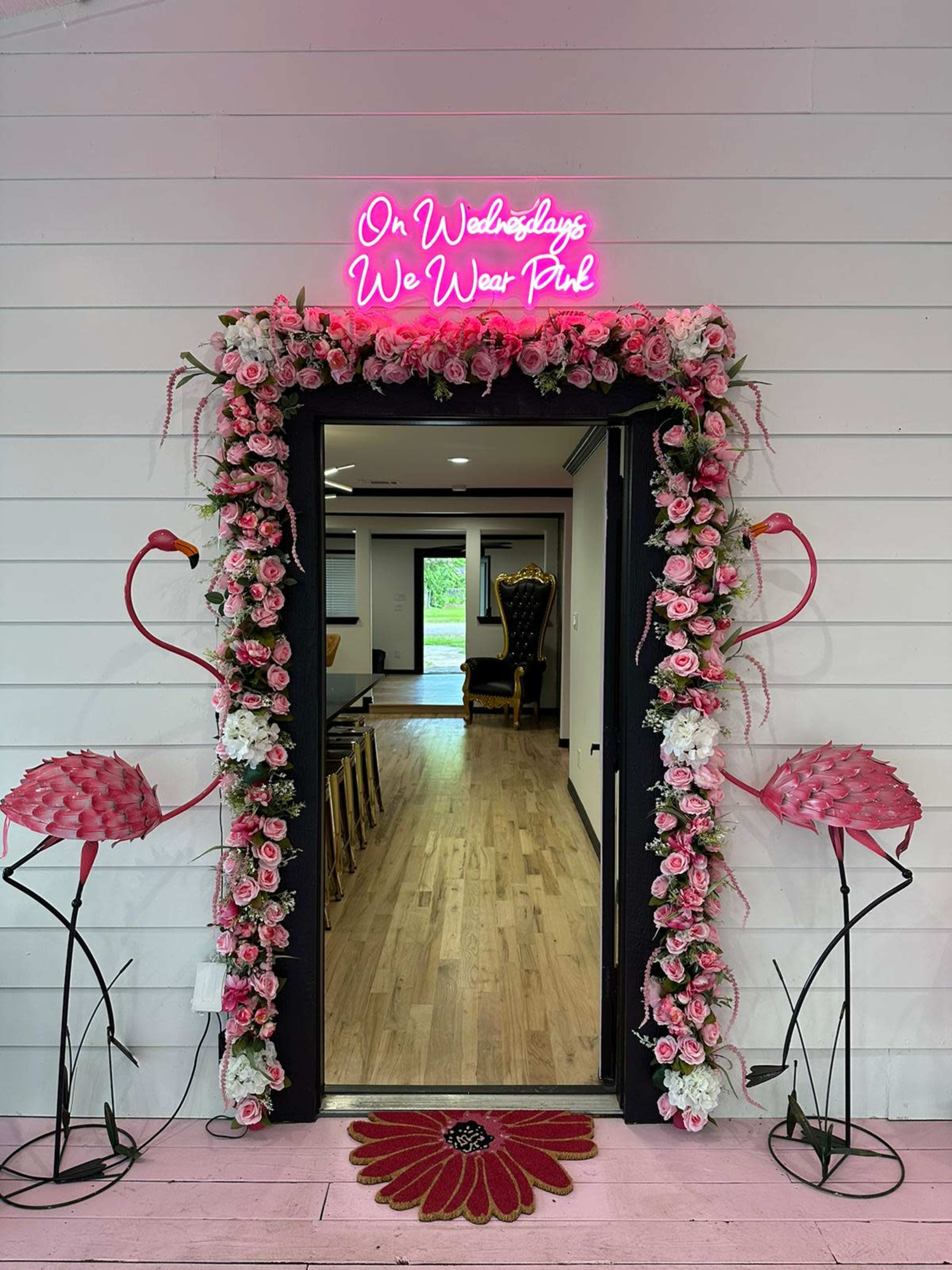 The entrance is adorned with a floral arch and pink flamingo sculptures, featuring a neon sign that reads, "On Wednesdays We Wear Pink."
