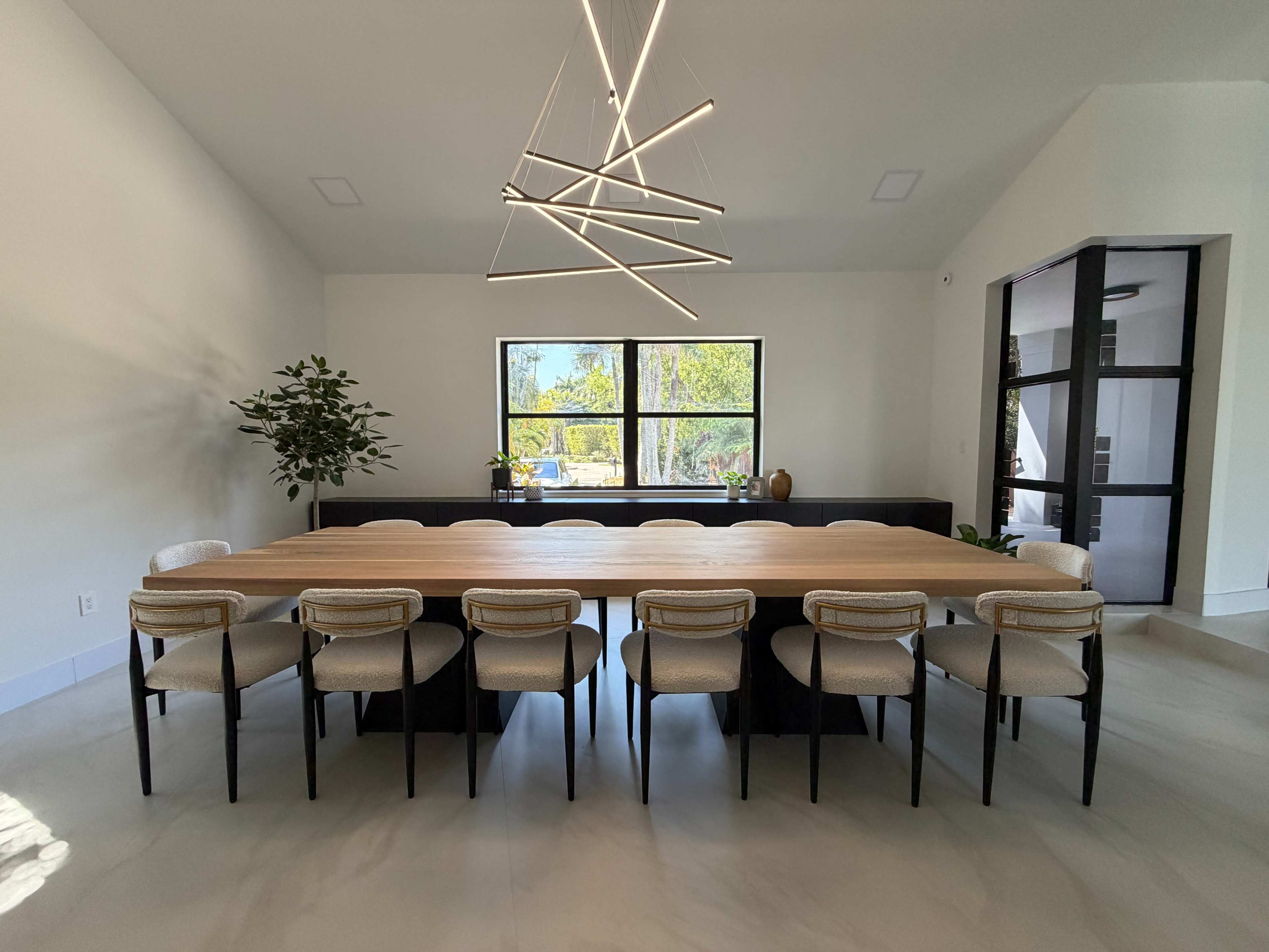 A long, rectangular dining table with chairs is set in a bright room featuring large windows and modern lighting.