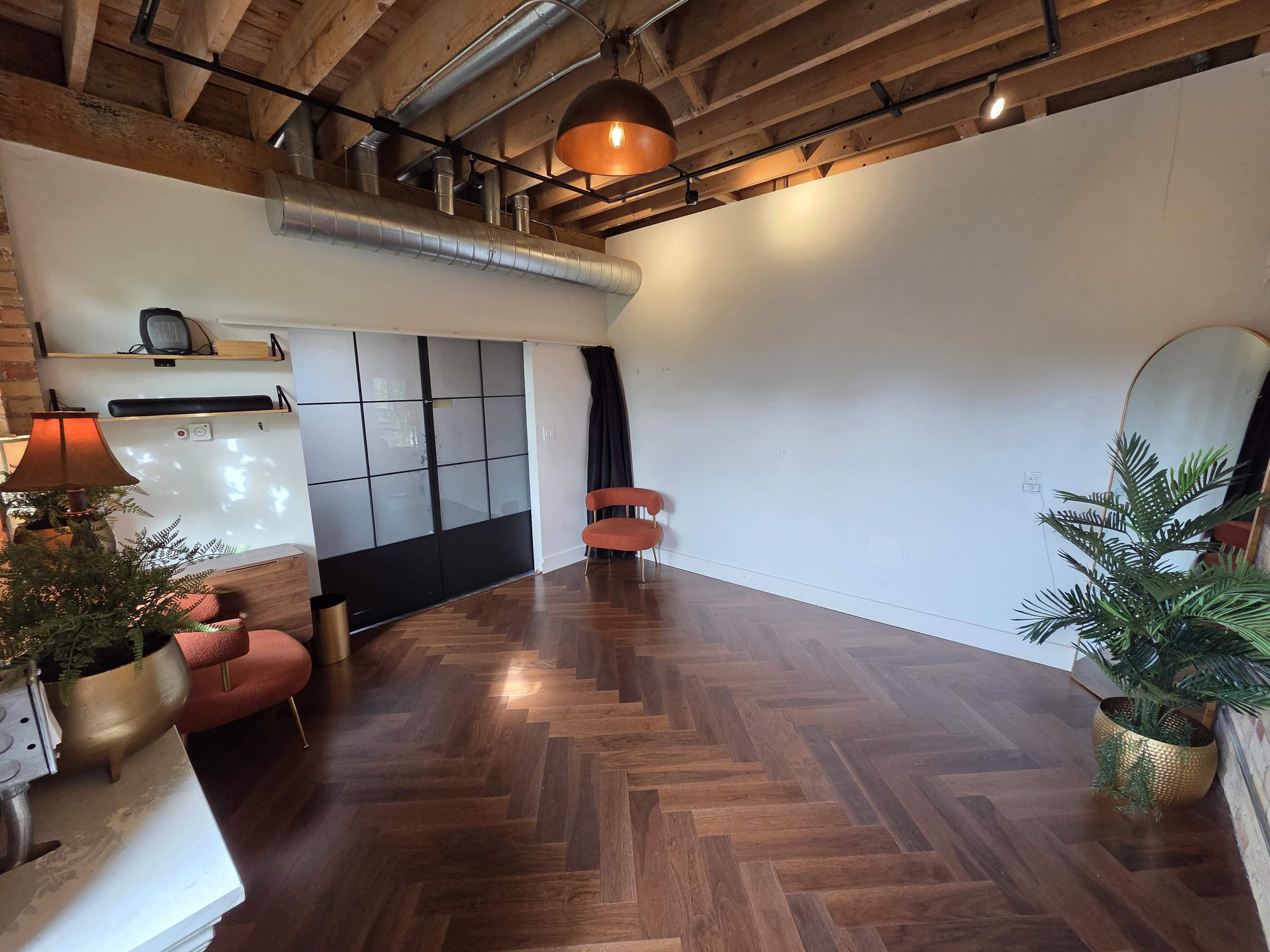 The image shows a spacious room with wooden flooring, exposed beams, and minimal furnishings, including two chairs and a plant, along with a large window and a dark curtain.