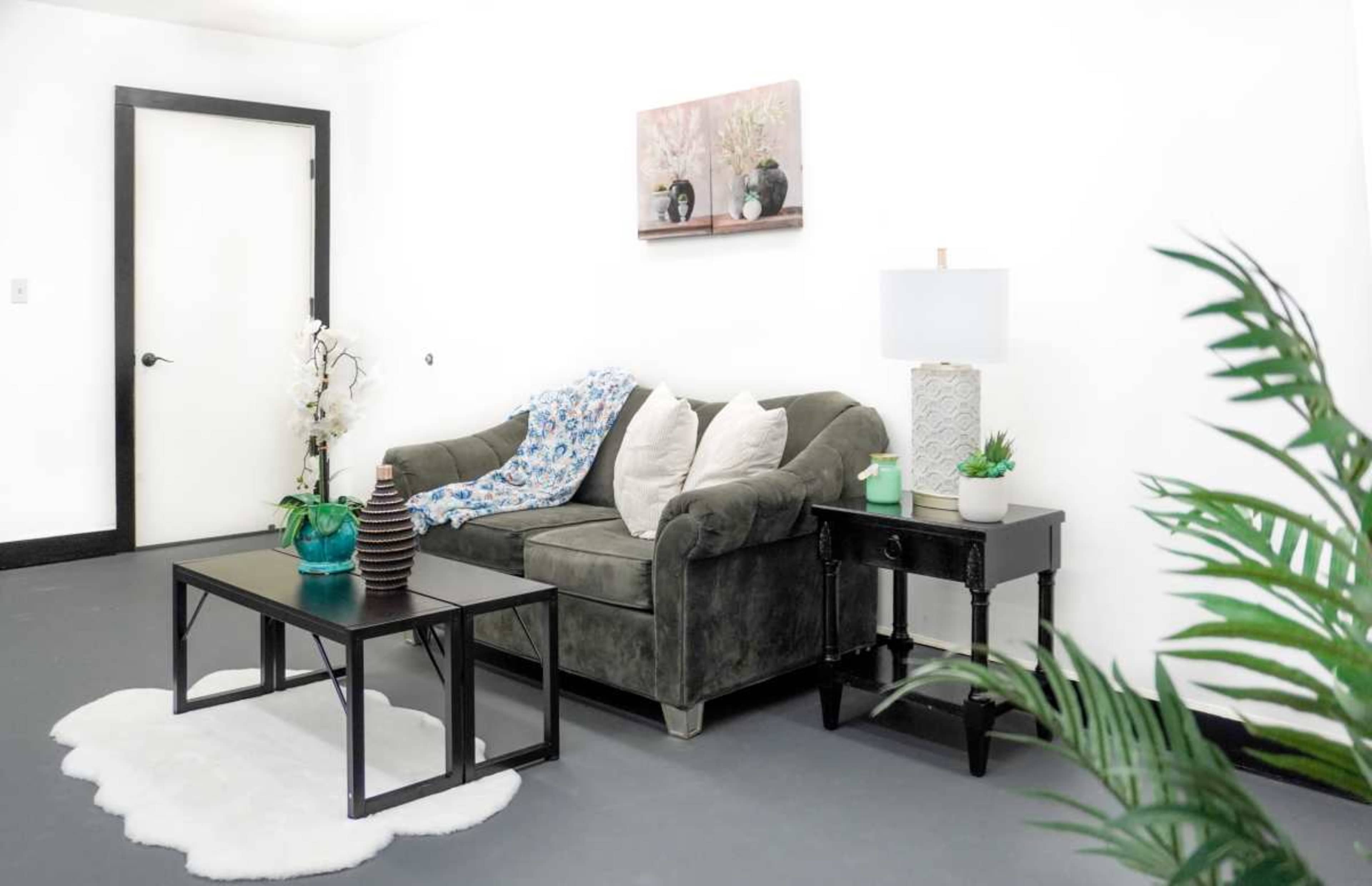 The image shows a living room scene with a gray couch, a coffee table, a lamp on a side table, and decorative plants, all set against a white wall.