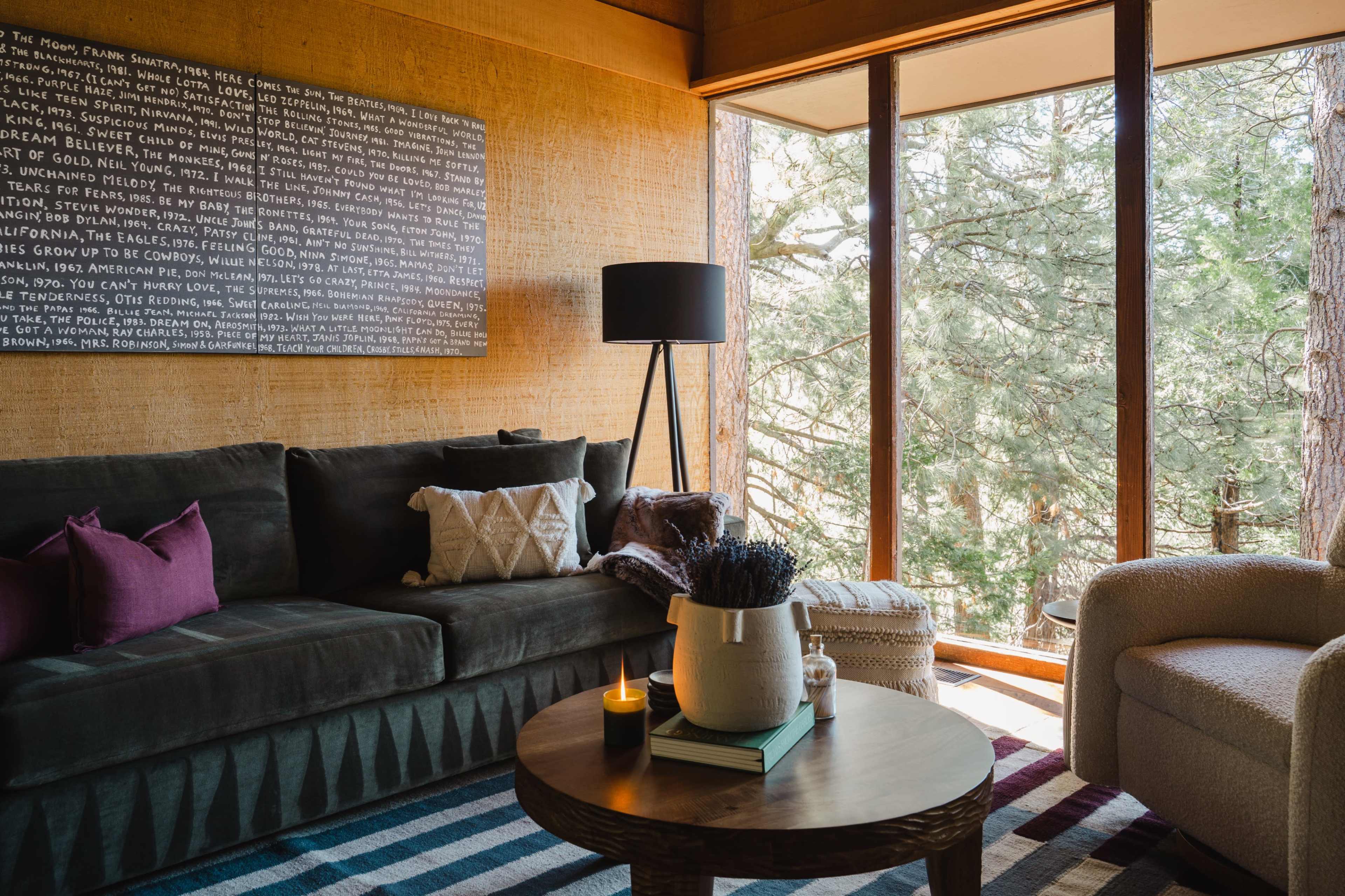 A cozy living room features a dark sofa, a round coffee table with a candle, and large windows overlooking trees.