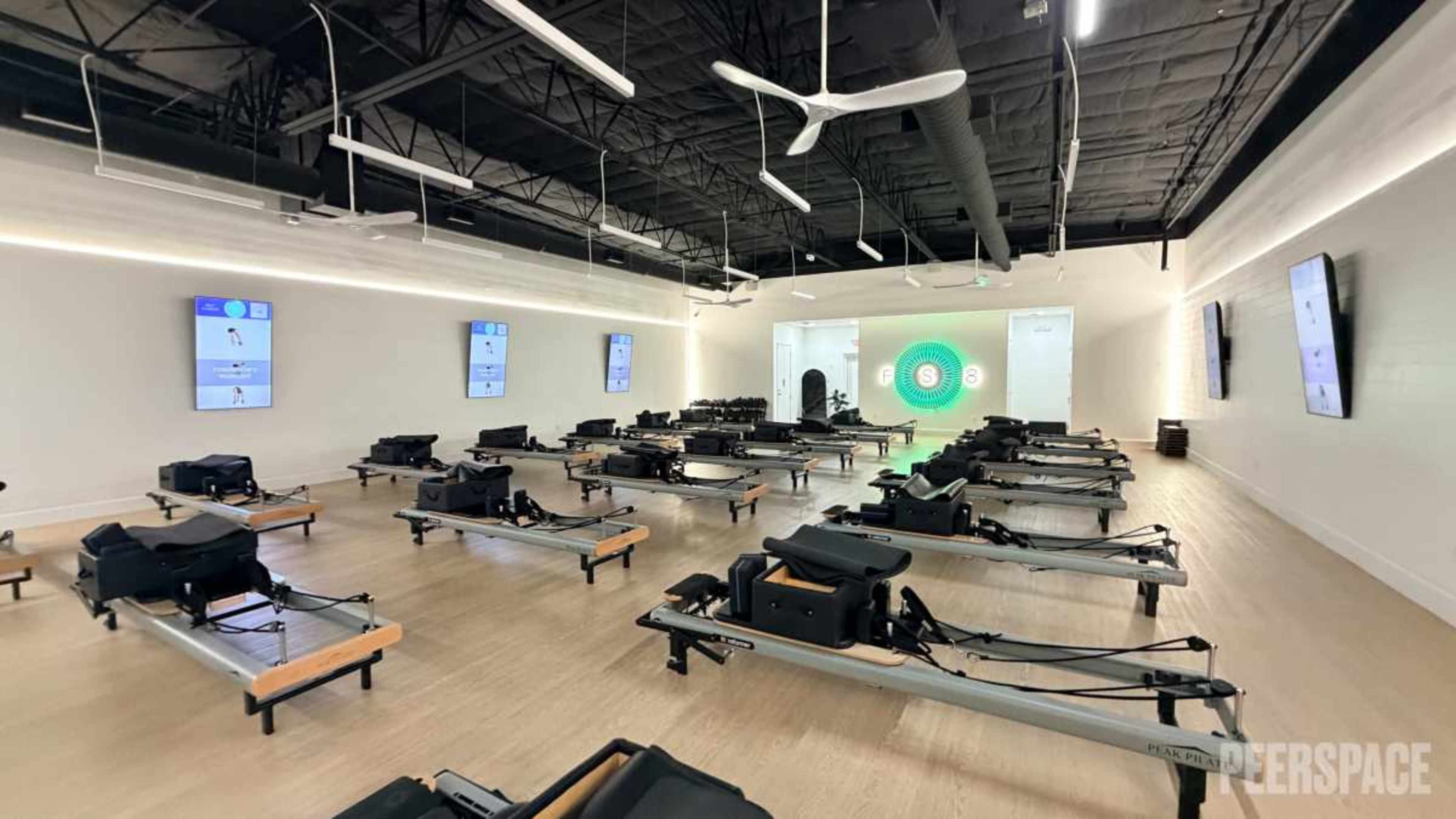 The image shows a spacious fitness studio equipped with multiple reformer pilates machines arranged neatly in rows, facing screens on the walls.