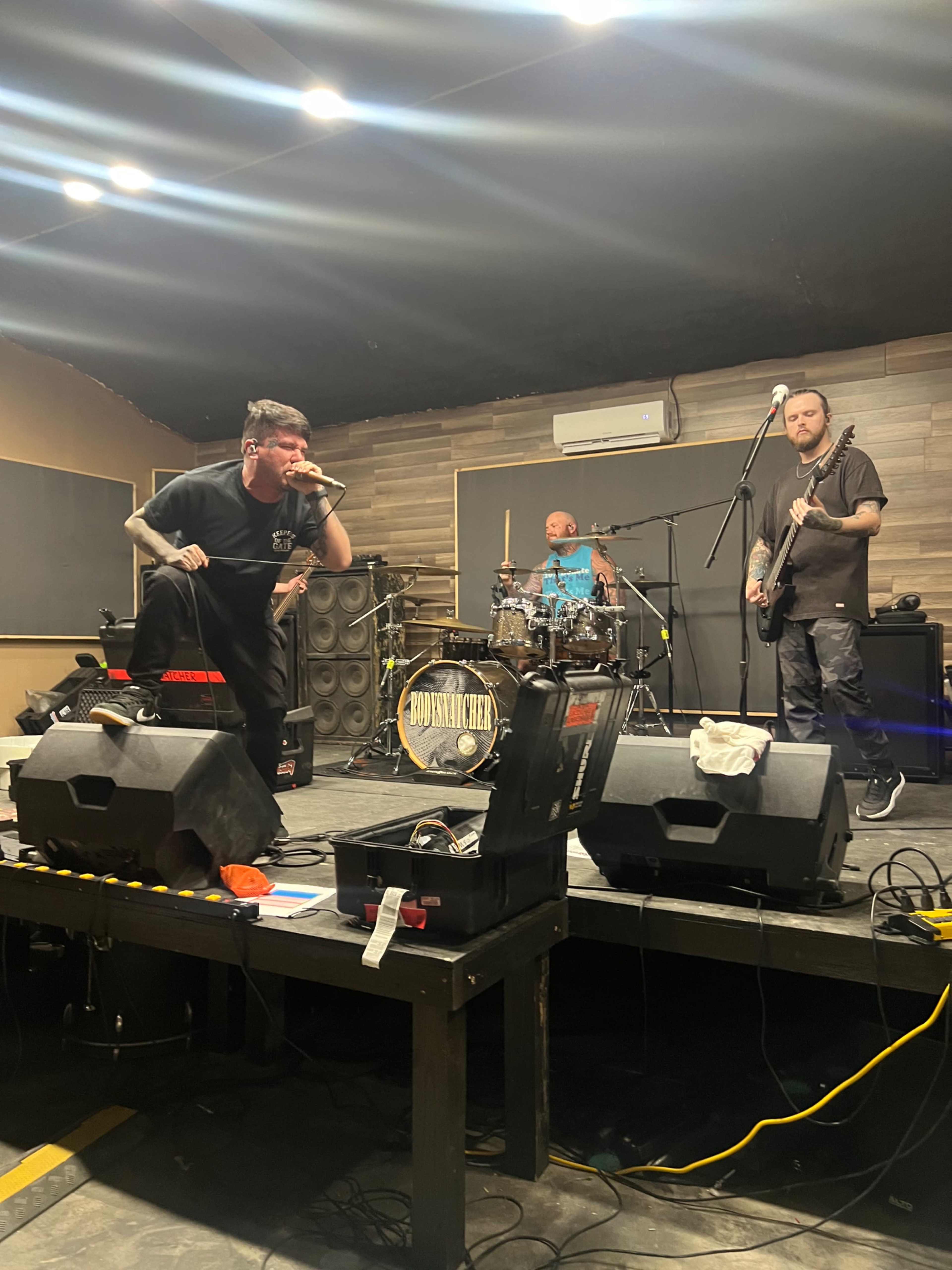 A band performs on a stage equipped with instruments and sound equipment, with a microphone in front of the lead singer, and other members playing drums and guitar.