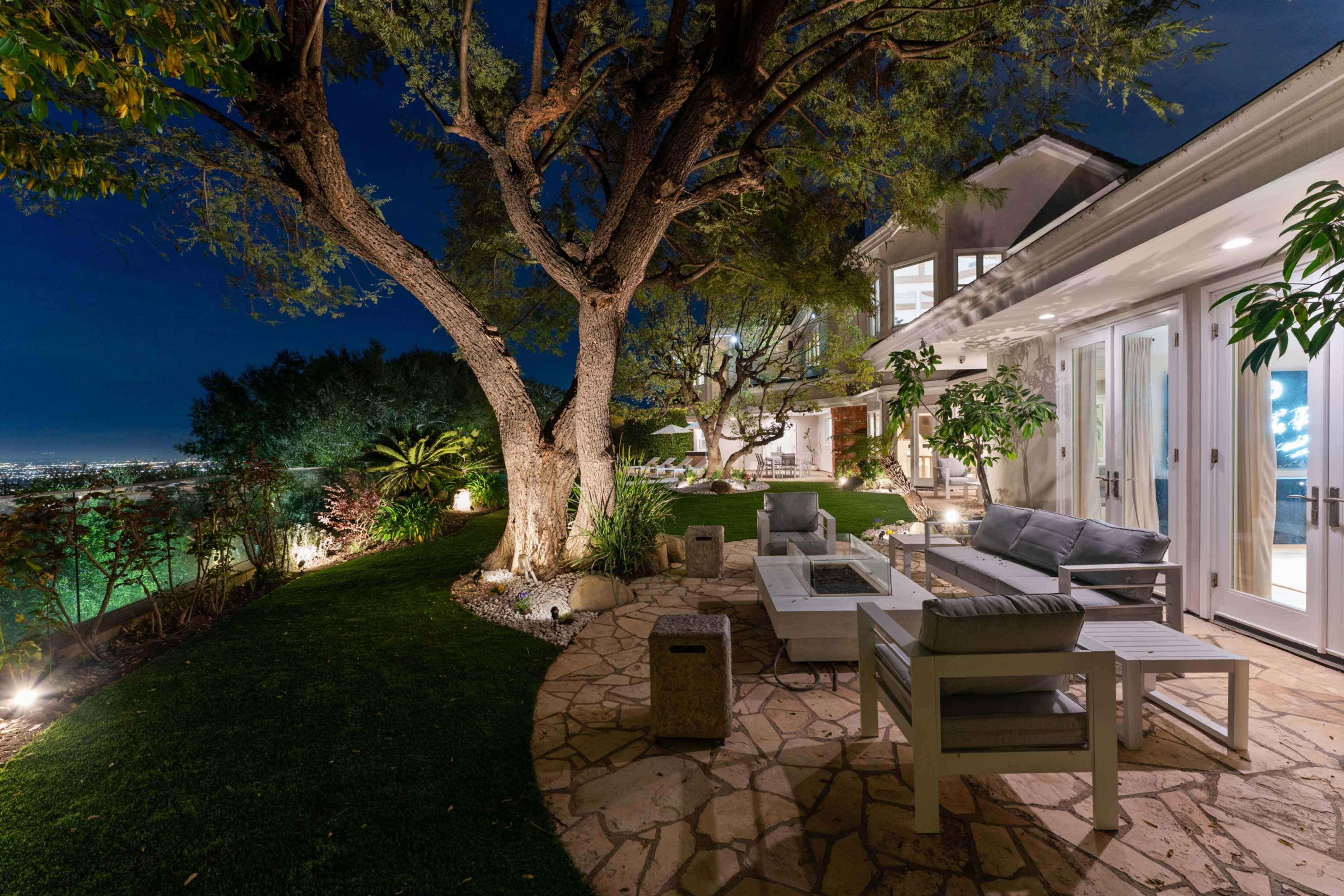 An outdoor patio features comfortable seating, a fire pit, and a tree beside a house, with city lights visible in the distance at night.
