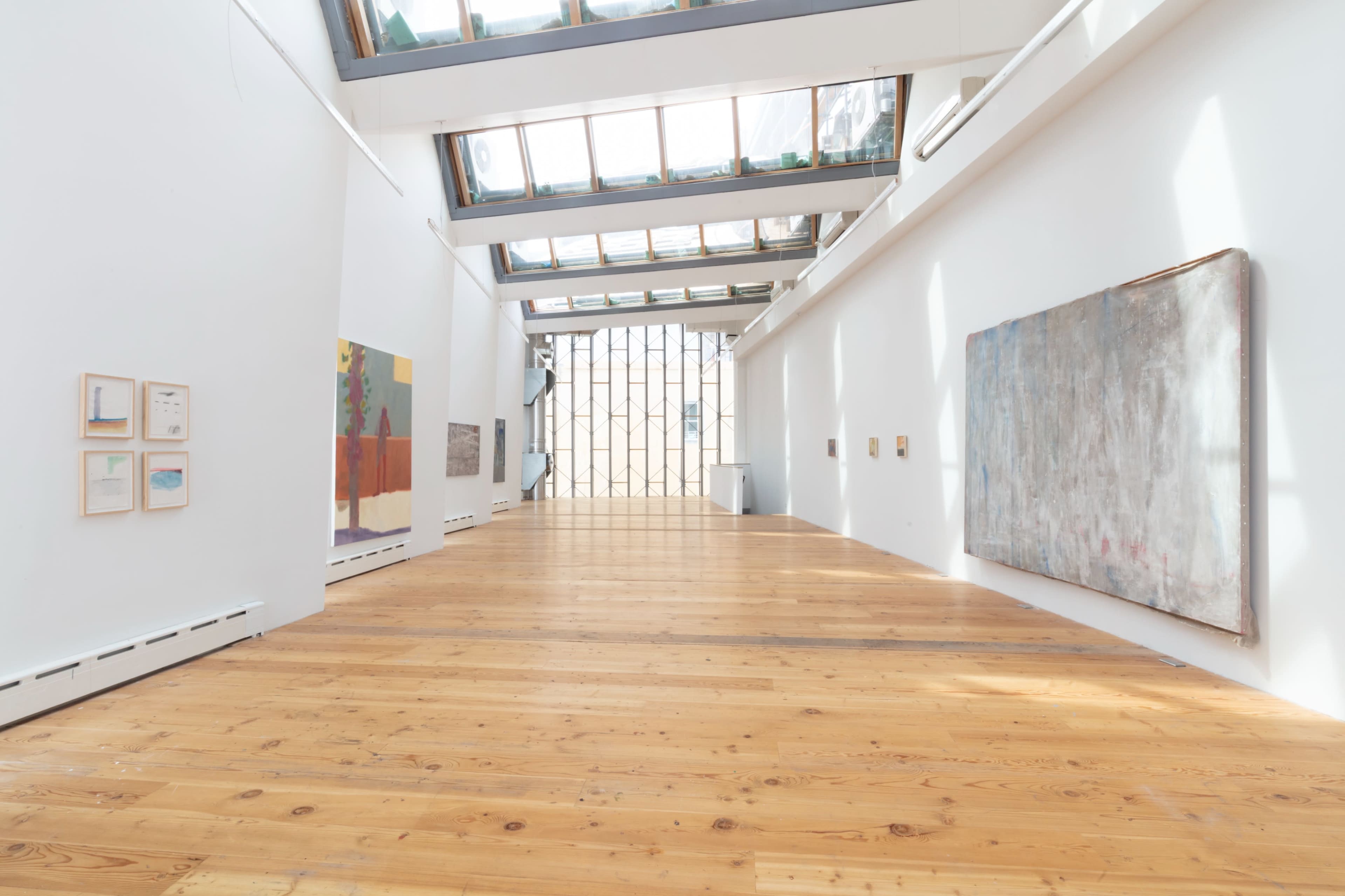 The image shows a bright, spacious art gallery with wooden floors and large windows, featuring several pieces of artwork on the walls.
