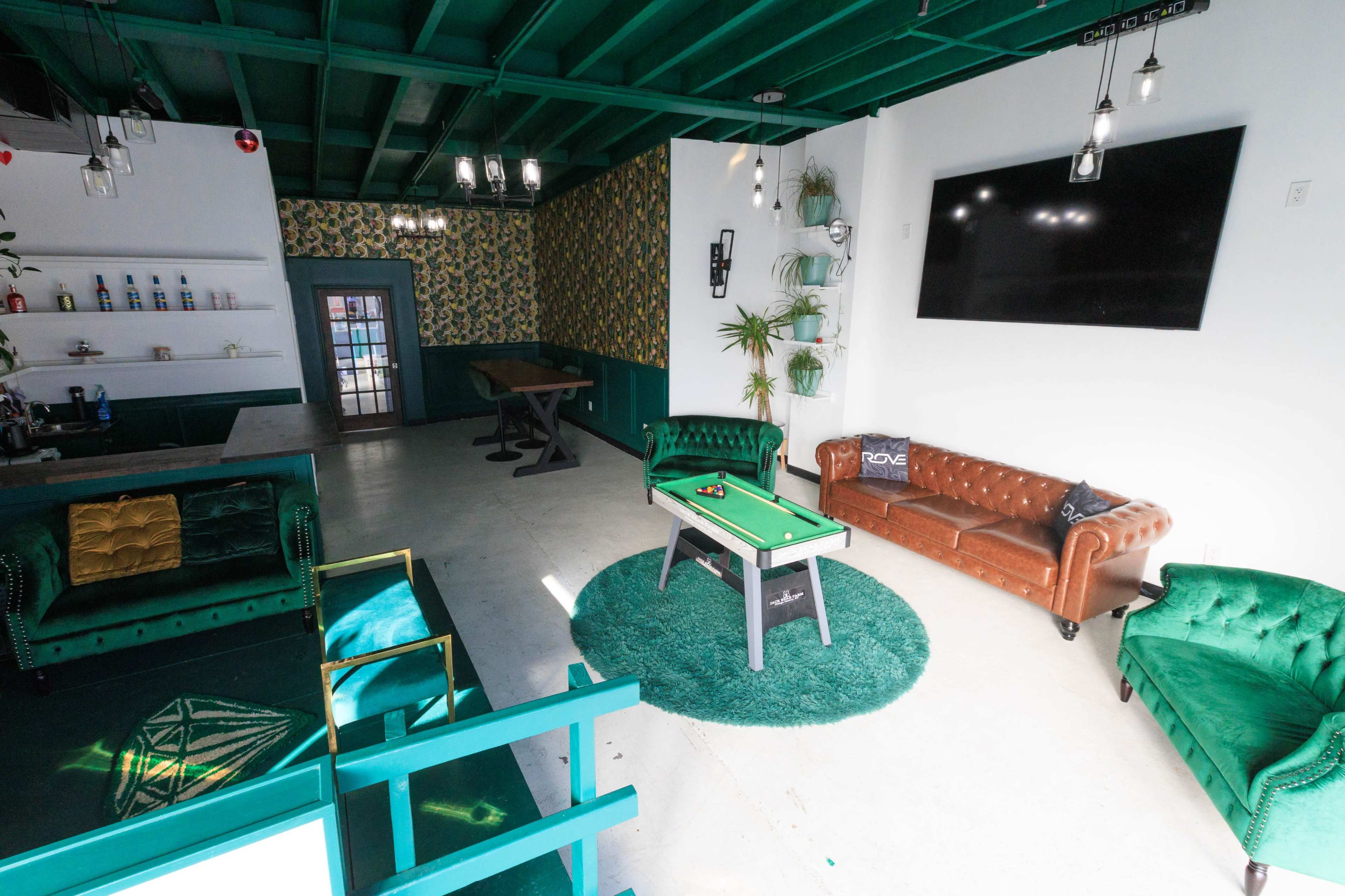 The image shows a contemporary lounge area featuring green and brown furniture, a decorative wall, and a small pool table on a circular rug.