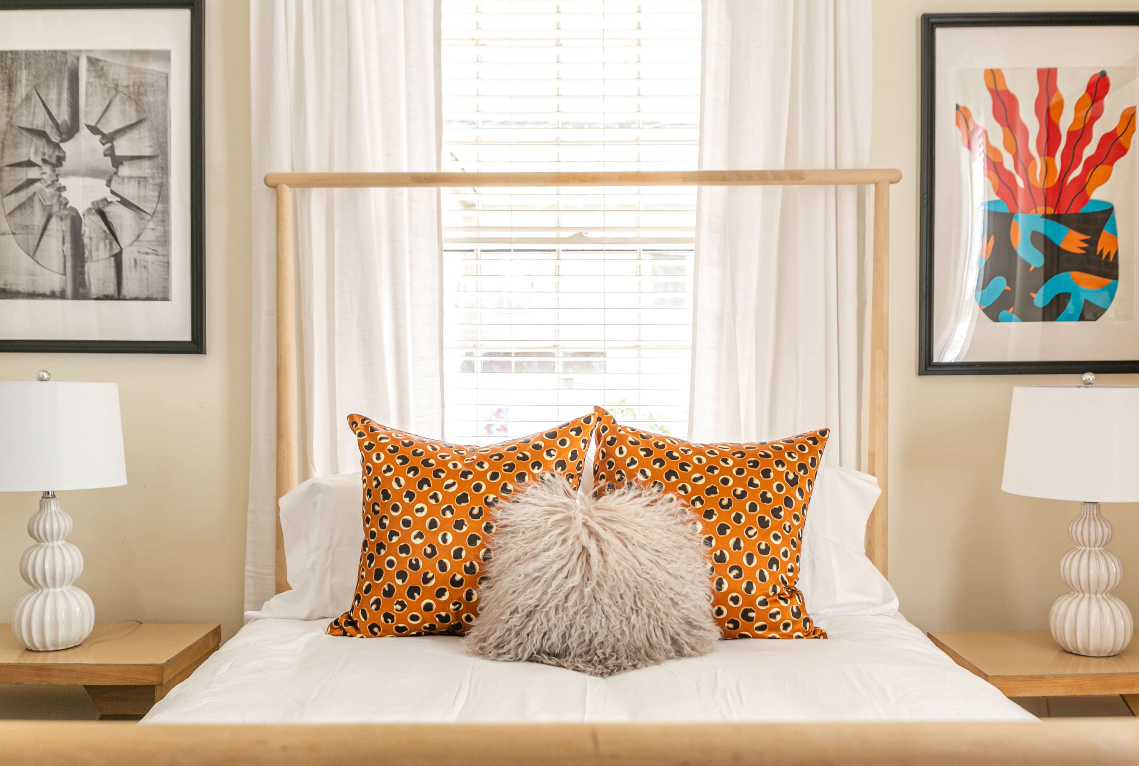 A neatly arranged bed with patterned orange and brown pillows, a fluffy center pillow, and framed artwork on the walls beside the window.