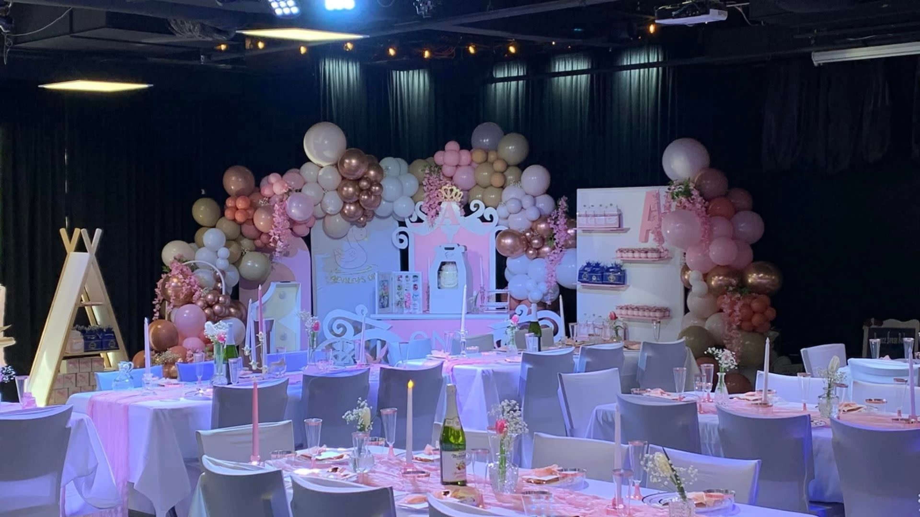 The image shows a decorated event space with tables set for a celebration, featuring balloon arrangements and a pastel-colored backdrop.