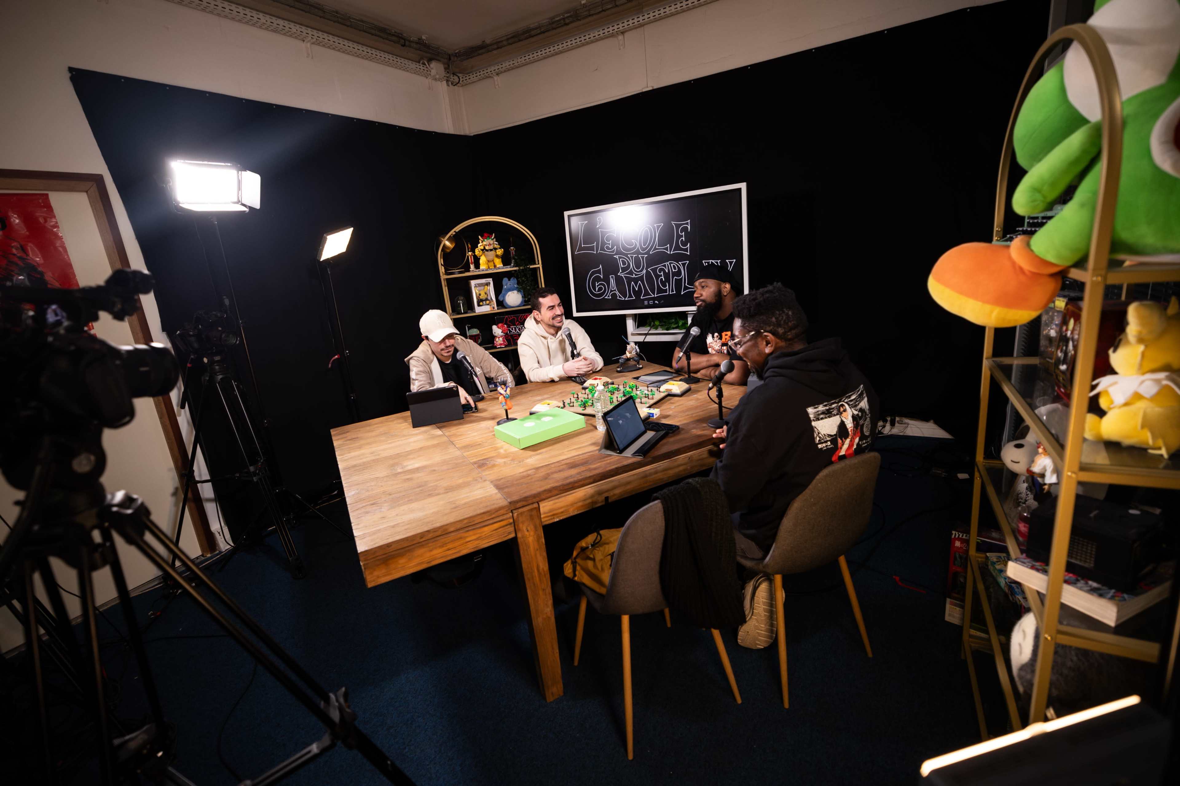 A group of four individuals sits around a wooden table equipped with devices for recording, surrounded by gaming merchandise and plush toys on shelves.
