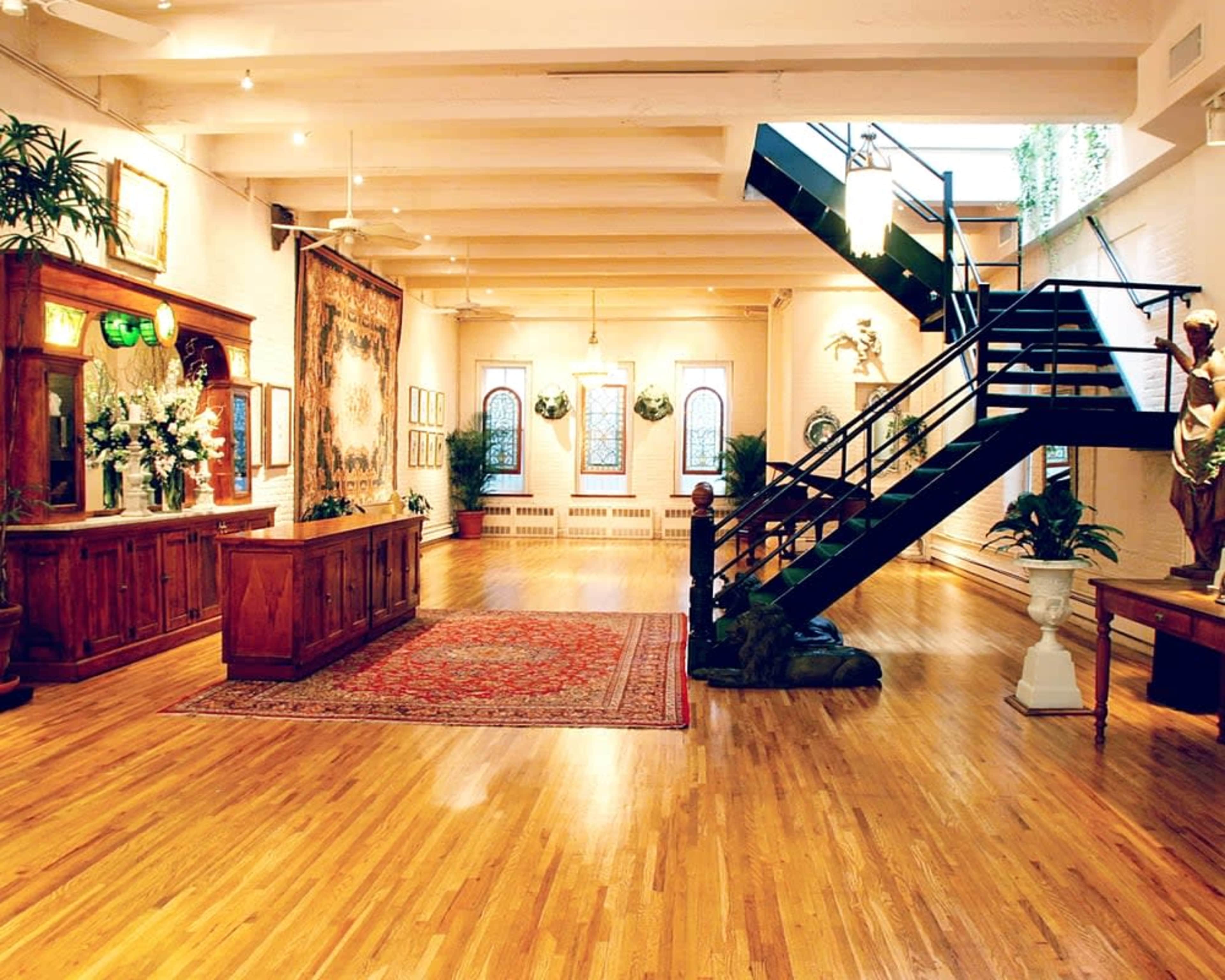 A spacious art gallery with wooden floors, a decorative rug, large windows, and a staircase leading to an upper level.