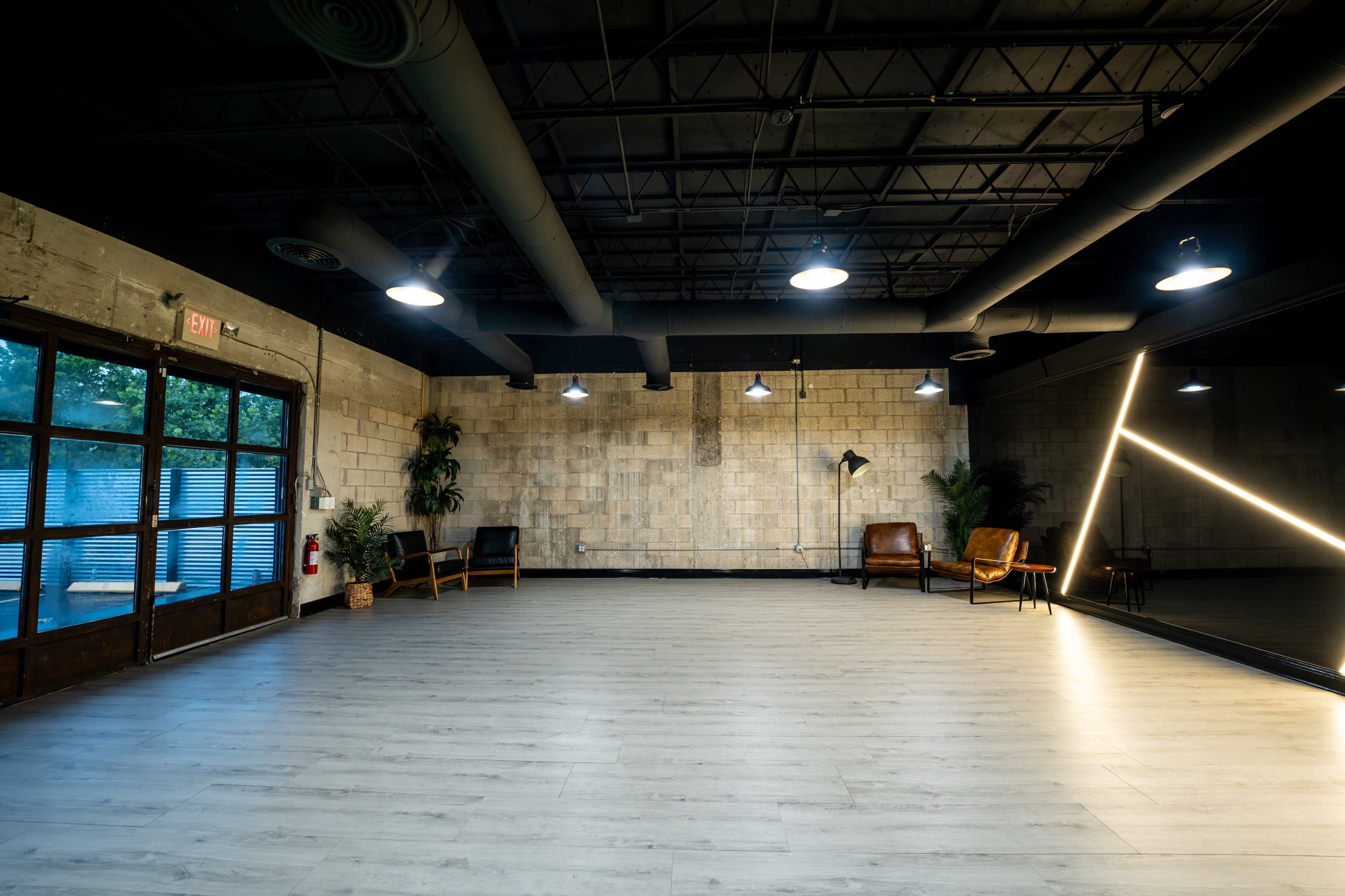 The image shows a spacious, well-lit studio with wooden flooring, gray walls, and a large mirror along one side, featuring two brown chairs and a potted plant.