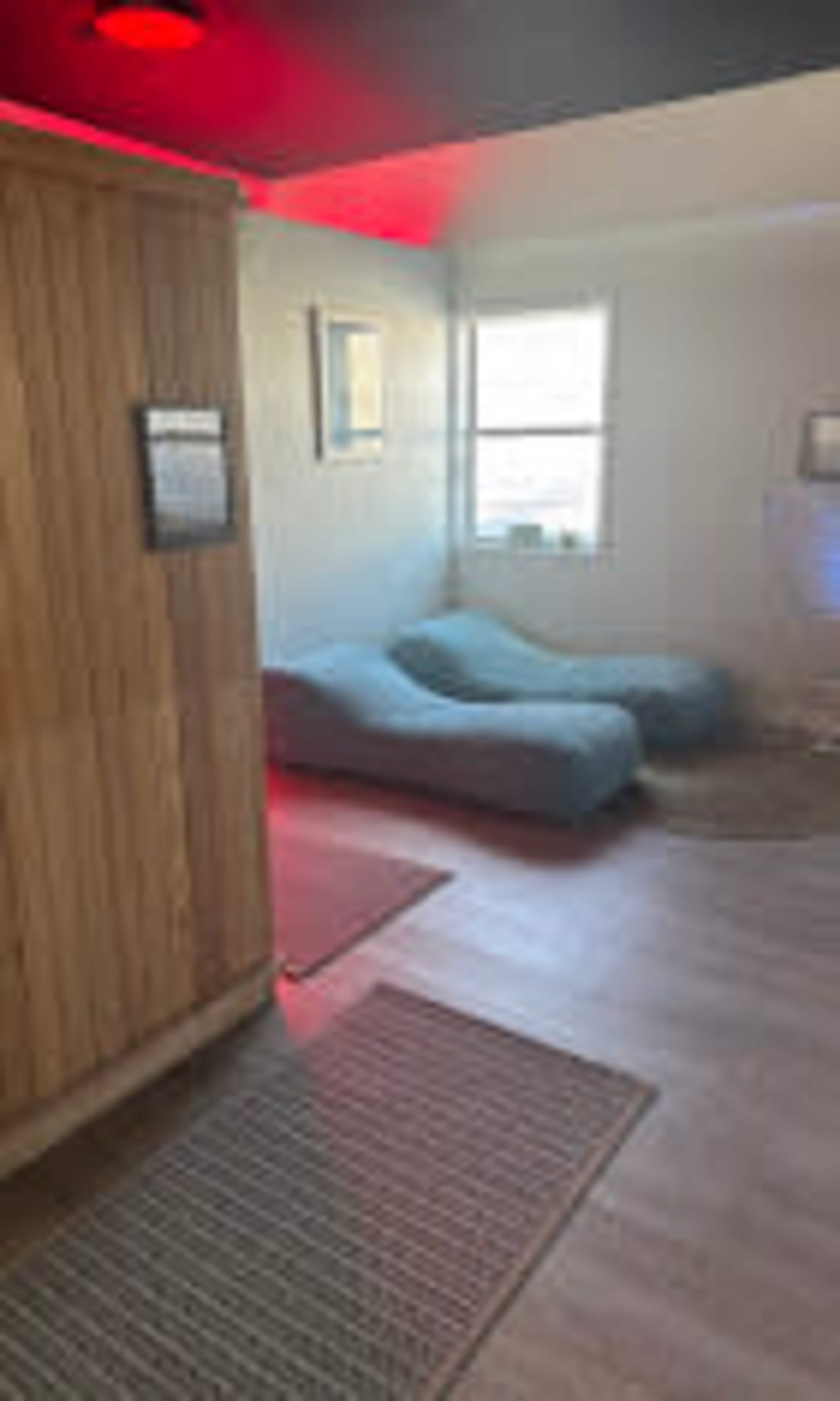 The image shows a room with two blue lounging chairs and a wooden wall, illuminated by red lighting and natural light from a window.