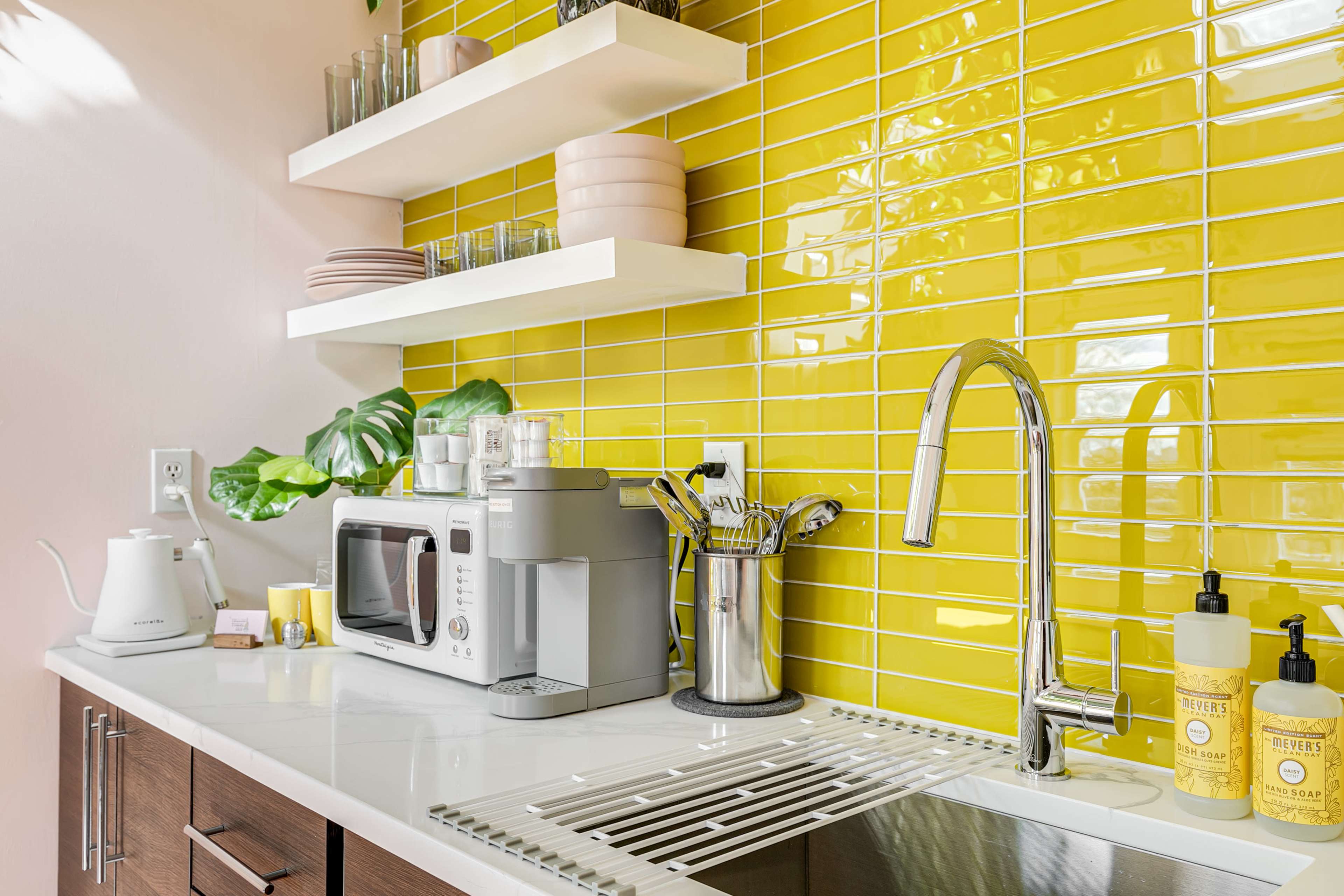 The image displays a modern kitchen with yellow tiled walls, showcasing a microwave, kettle, and various kitchen utensils on the countertop.