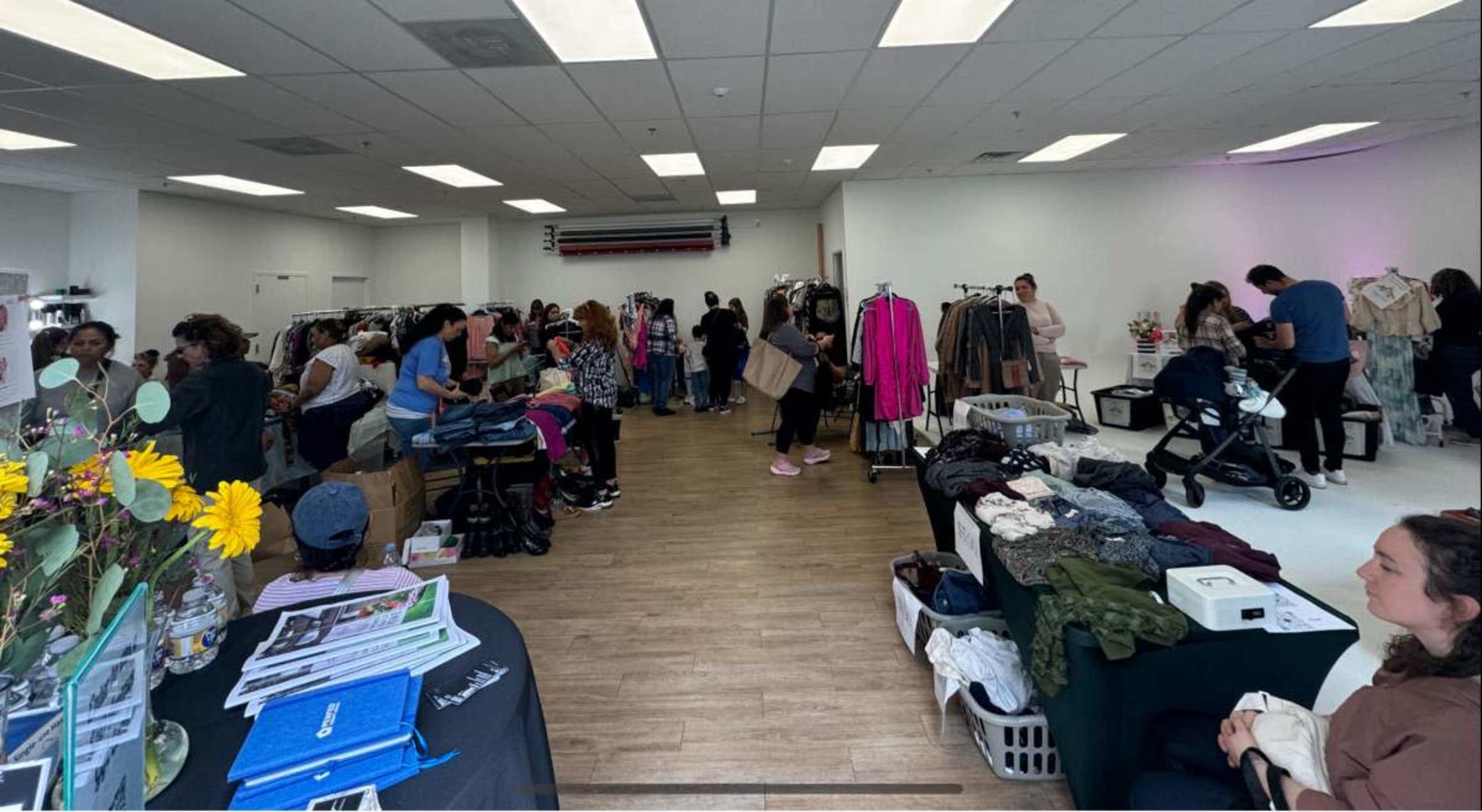 A clothing boutique filled with customers browsing various racks of clothing and accessories, while others are engaged in conversation or pushing strollers.