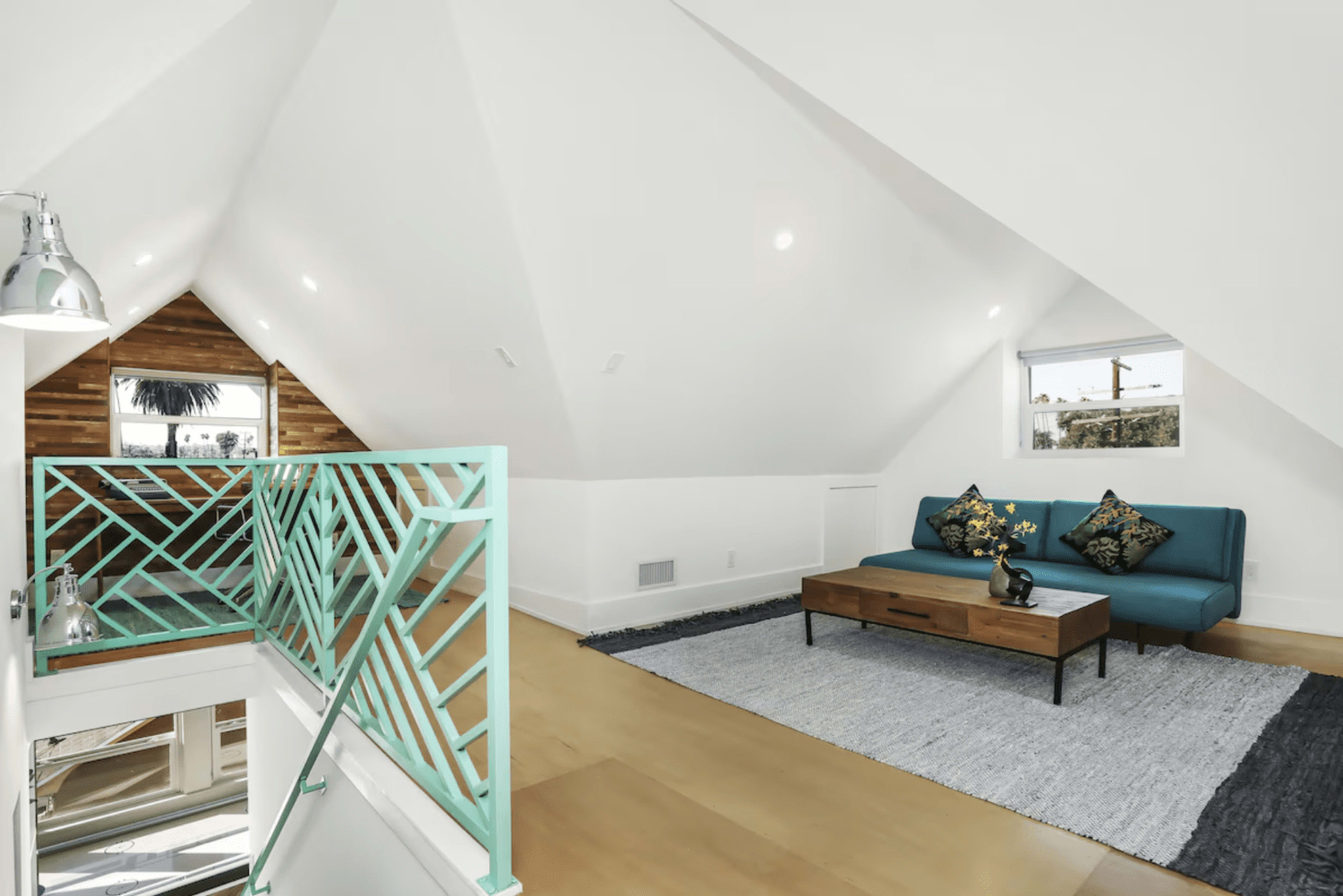 The image shows a bright, modern loft space with a teal sofa, a wooden coffee table, and a geometric railing overlooking an open area.