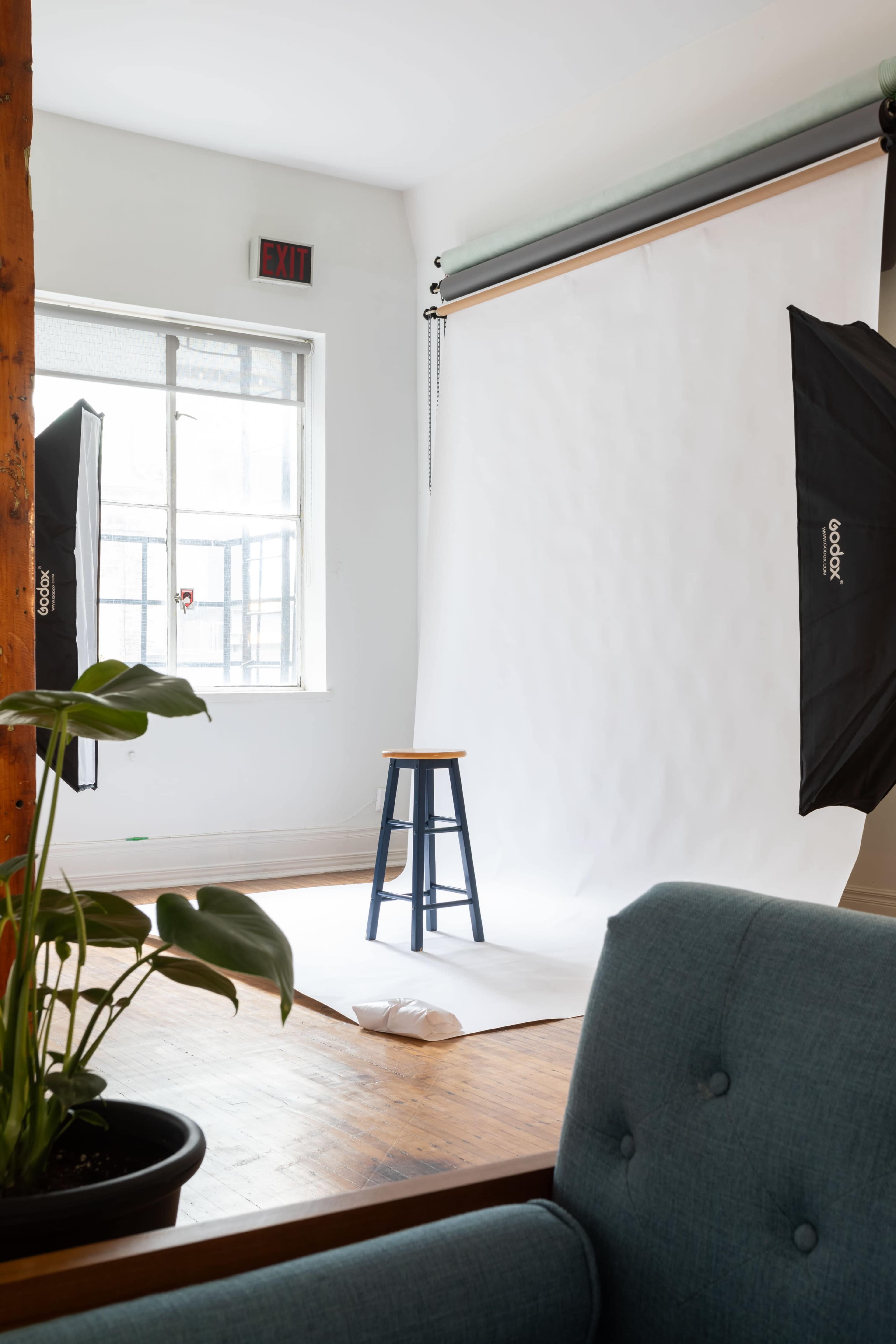 A photography setup with a blank backdrop, a stool, and studio lights in a room with a window.