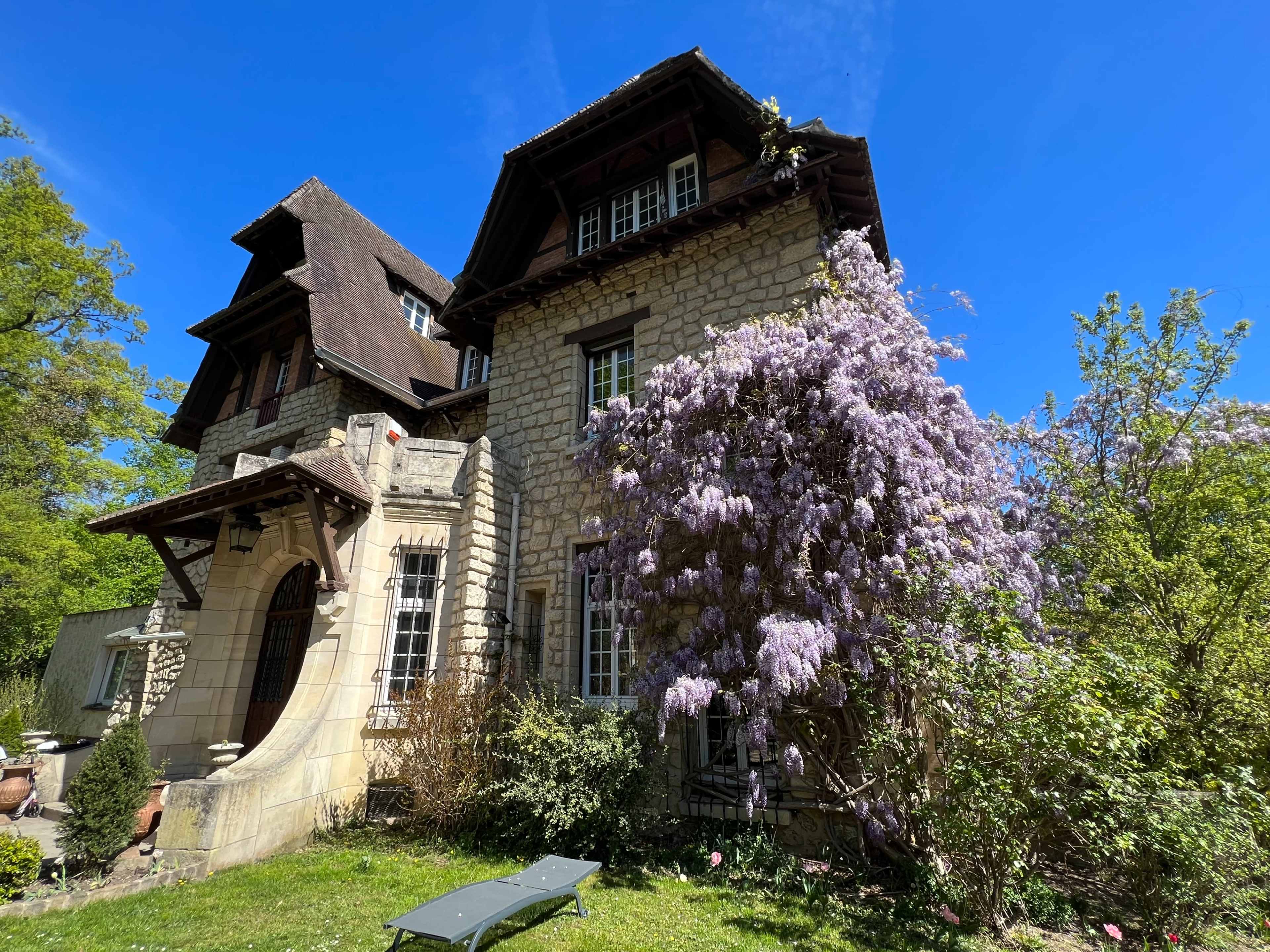 A stone house with a sloping roof is adorned with blooming wisteria and surrounded by green foliage under a clear blue sky.
