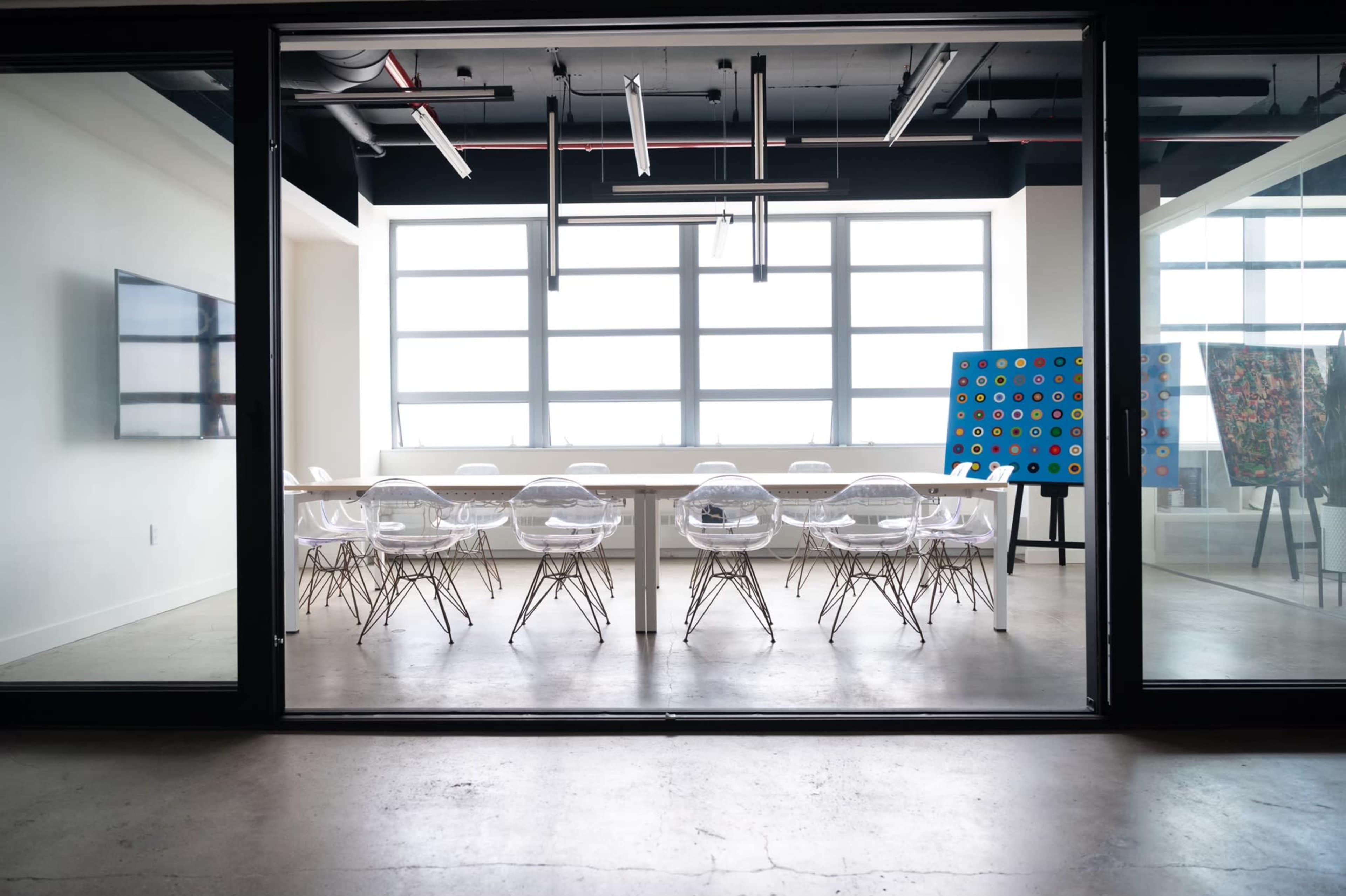 A modern conference room features a long table surrounded by transparent chairs, with large windows allowing natural light and artwork displayed nearby.
