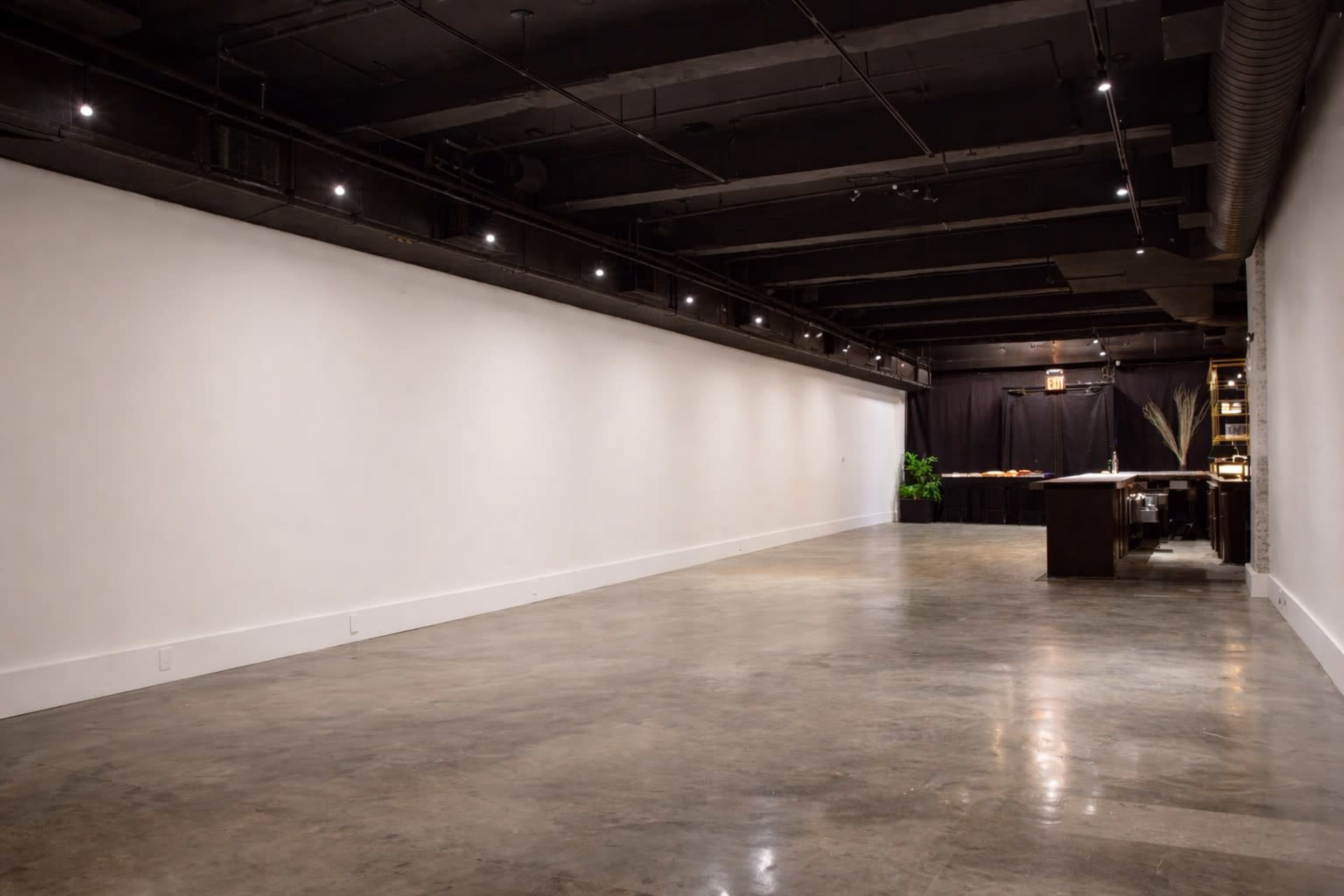 The image shows a large, empty room with polished concrete floors, white walls, and a dark backdrop at one end.
