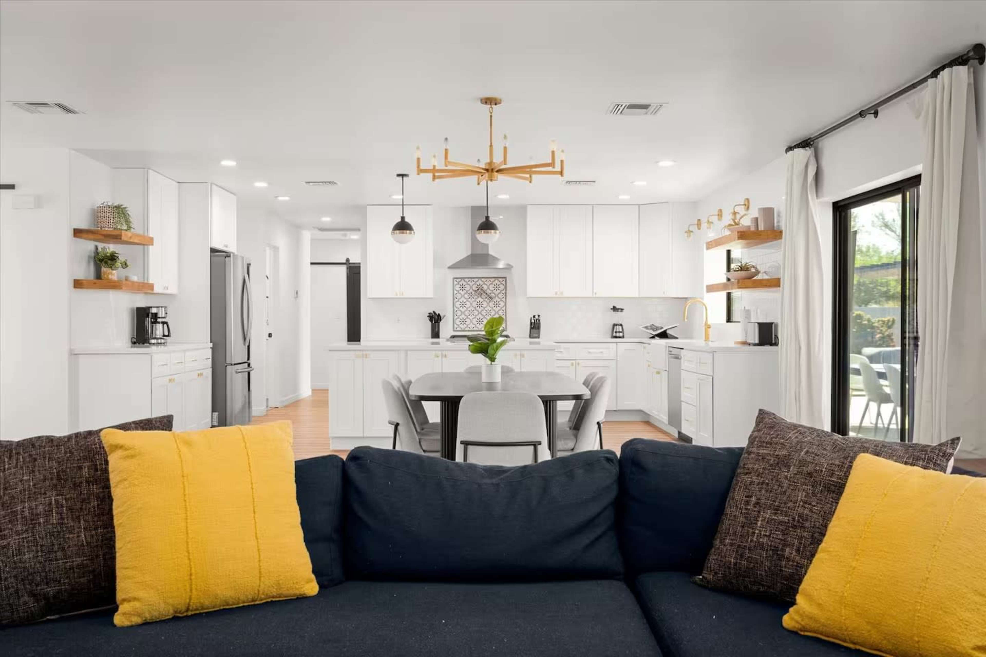 A modern kitchen and dining area is visible, featuring white cabinets, a gray island, and a table surrounded by six chairs, with a sofa in the foreground adorned with yellow and gray pillows.