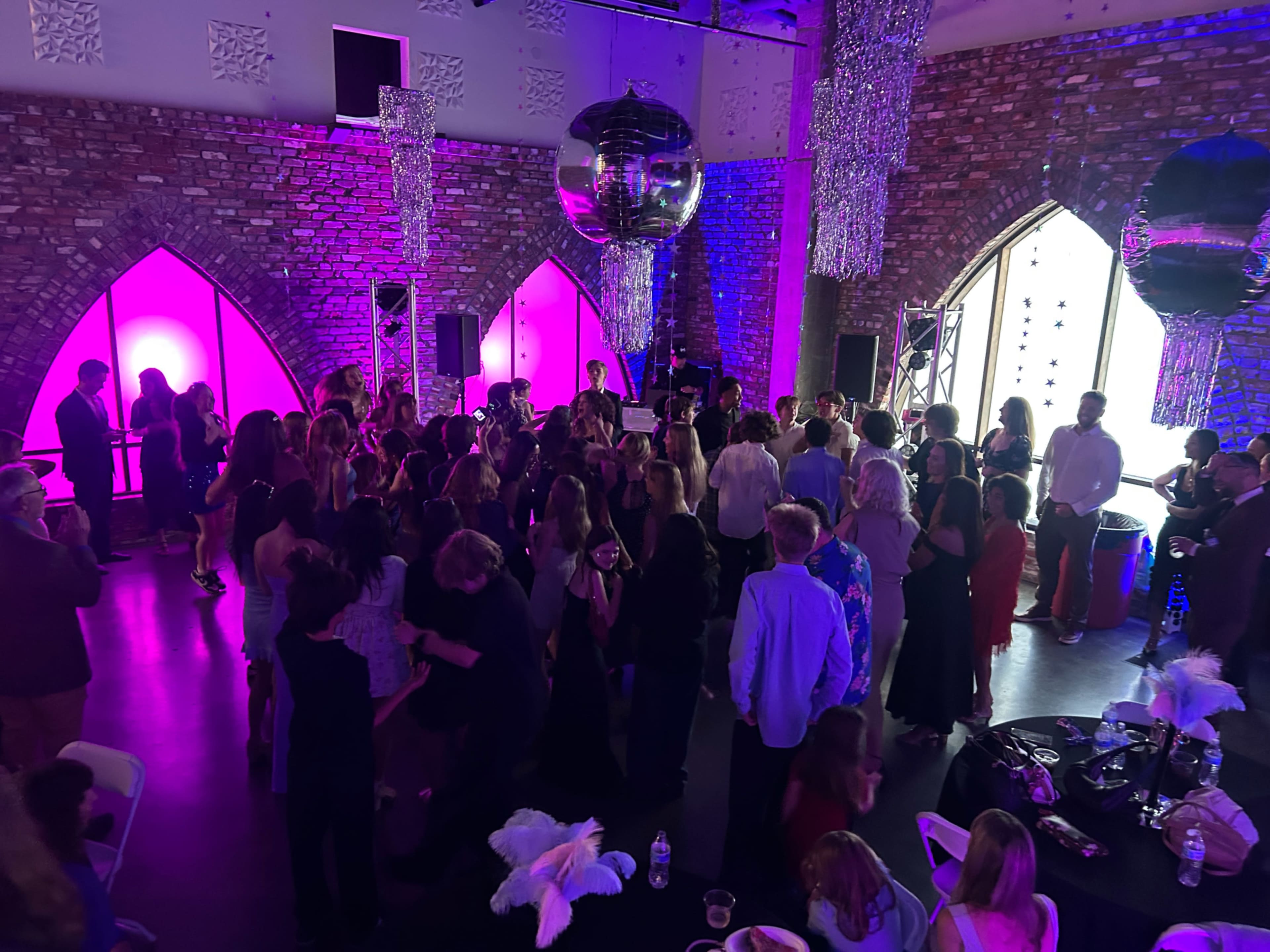 A crowded event space features guests dancing under large disco balls and colorful lighting.