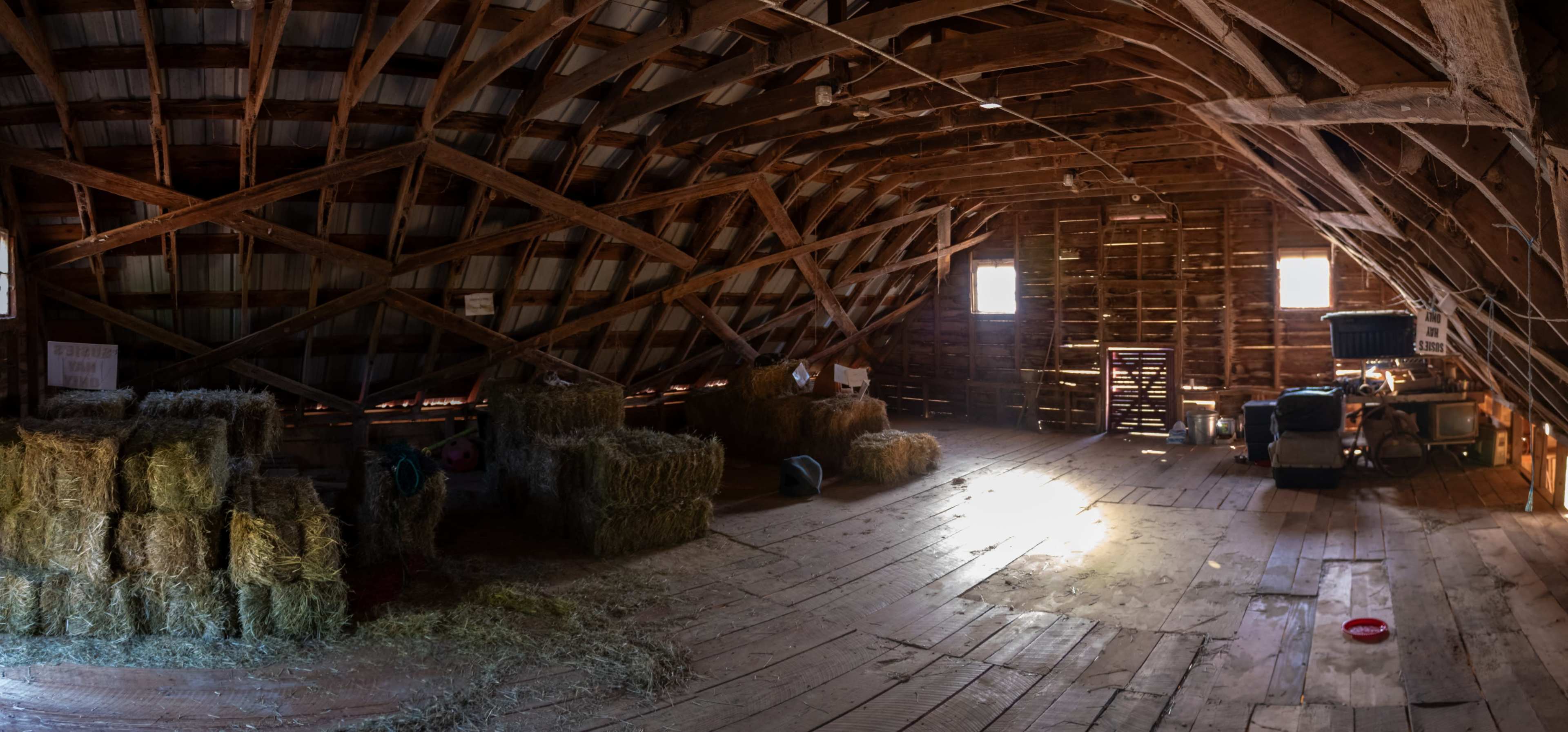 The image shows the interior of a rustic barn with wooden beams, scattered bales of hay, and a few windows allowing light to enter.