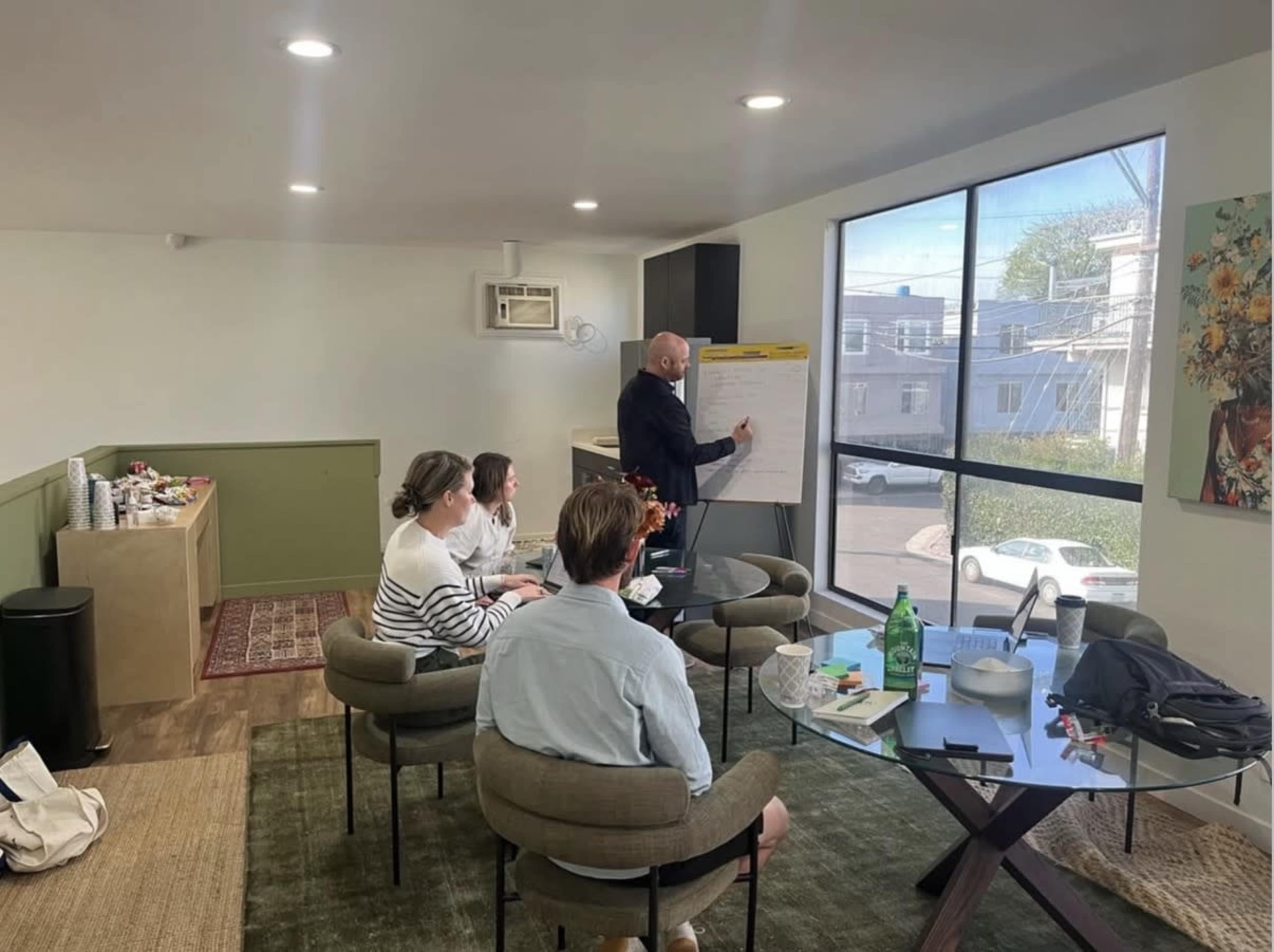A group of three people sit around a table while a fourth person stands and presents ideas on a flip chart in a well-lit room with large windows.